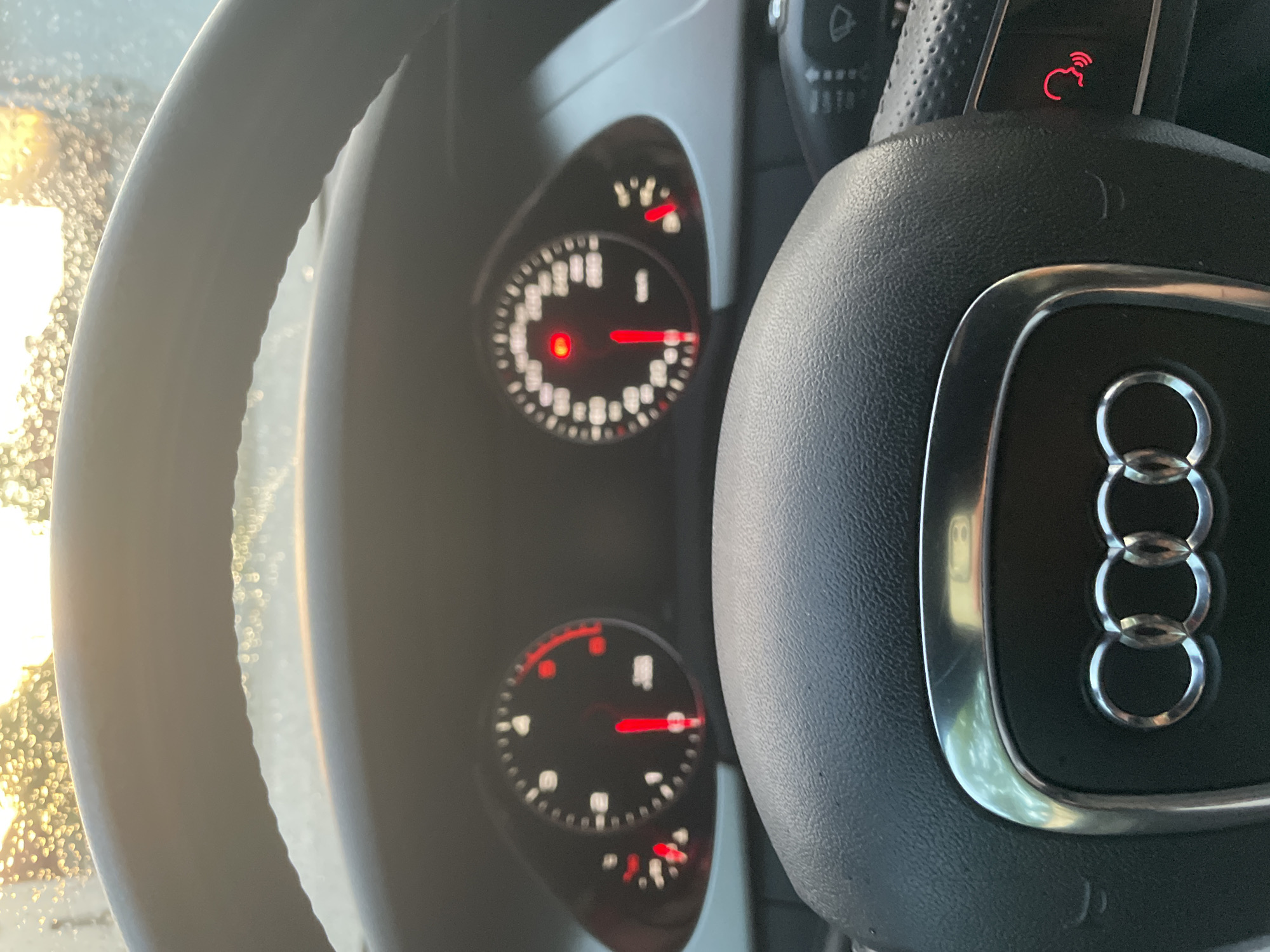 The image displays the interior of an Audi car, likely in Ihtiman, Bulgaria, viewed from the driver's perspective. Bright sunlight streams in from the left, illuminating the dashboard and a portion of the black leather steering wheel, and creating a bokeh effect from what appears to be water droplets or condensation on the windshield.
The prominent chrome Audi four-ring logo is centered on the steering wheel. Above it, on the right spoke, a red illuminated icon of a head with sound waves suggests an active voice control or speech input function.
Behind the steering wheel, two circular gauges are visible on the dashboard. The upper gauge shows a red needle pointing to a low value, possibly indicating zero speed or RPM. This gauge also displays a small red warning light at the top right and a visible "P" symbol, indicating the car is in Park. The lower gauge also features a red needle pointing to a low reading, accompanied by smaller red indicators at its bottom right. The illuminated red elements on the dashboard suggest the car is either stationary with the engine off, in accessory mode, or has just been started. The overall scene depicts a bright day, possibly following rain or during the early morning.