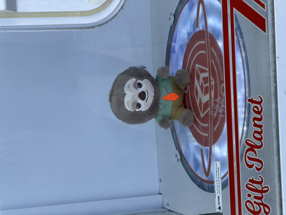 An interior view depicts a single plush toy situated within a transparent arcade prize machine. The toy is a brown, furry creature, approximately 20-25 cm in height, resembling a sloth, with a white face, dark nose, and embroidered eyes that appear partially closed with a slight smile. It is dressed in a light green striped collared shirt, khaki pants, and a prominent orange tie. The toy sits upright on a circular, reflective platform within the machine. This platform is illuminated from below, casting a bluish light, and features a central red graphic resembling a planet or orb with concentric rings and a stylized cubic shape in its core. The lower front panel of the machine is silver-toned, bearing the words "Gift Planet" in red script. A black and white warning label on the platform's edge states: "CAUTION! Do not push or lean on the glass to avoid glass breaking damage and danger." The interior walls of the machine appear light blue-grey. The scene is located within a "Gift Planet" establishment in Rishon LeZion, Israel, consistent with an arcade or entertainment venue. No human subjects are visible.