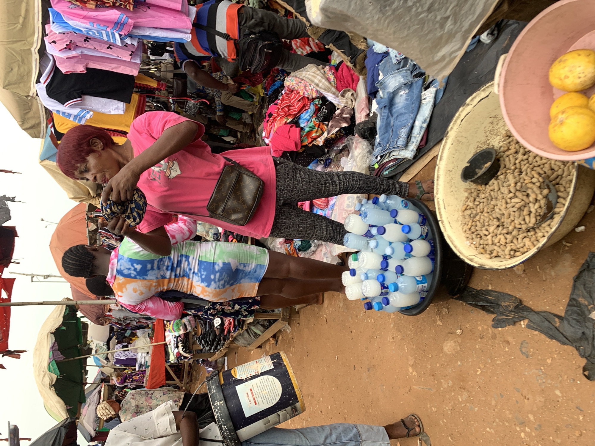 This image captures a vibrant street market scene in Nyanya, Nigeria. The focus is on several individuals interacting with a variety of goods. In the foreground, a woman with short red hair, wearing a pink t-shirt and patterned leggings, is examining something in her hands. She has a crossbody bag with a Louis Vuitton pattern. Beside her, a woman with braided hair wears a colorful, tie-dye dress and appears to be looking down.
The market is stocked with an abundance of clothing items, including stacks of t-shirts and folded garments. There are also several plastic bottles of water arranged in a tub, and a larger container filled with peanuts. In the background, more market stalls and people are visible, suggesting a bustling environment. The ground appears to be sandy or dirt, consistent with an outdoor market.
Notable details include the bright sunlight, indicating it is daytime. The overall atmosphere seems busy and active, typical of a marketplace. The image provides a glimpse into daily life and commerce in the region. There is a visible "CAUTION" label on a dark blue and white drum in the lower part of the image.
