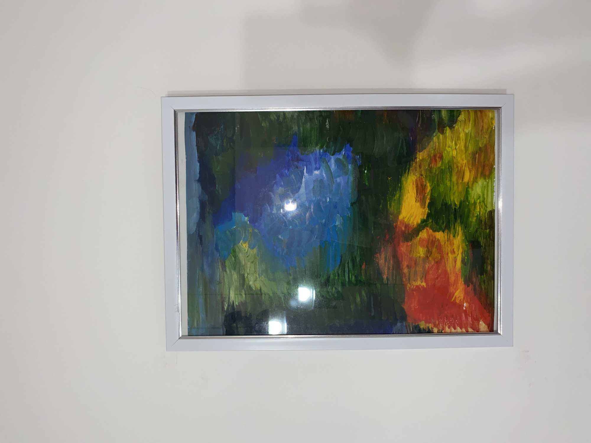 An abstract painting in a rectangular frame hangs on a plain, light-colored wall. The artwork features dynamic, broad brushstrokes in a vivid color palette. Dominant hues include deep blues and various greens towards the center and left, transitioning to bright yellows and reds on the right side. The painting's style is abstract, with no identifiable figures, objects, or landscapes.
The painting is protected by glass, which shows prominent reflections, particularly two bright, circular light sources near the bottom center, indicating an indoor setting with artificial lighting. The frame is a simple, modern design, light grey or silver in color, and creates subtle shadows on the wall. The wall itself is a uniform, light neutral shade, likely off-white or cream. Some faint imperfections, such as a thin dark line or minor smudges, are visible on the wall in close-up views.
The scene is static, focusing solely on the displayed artwork. No people are present, and no specific activity or event is taking place. There is no visible text. The overall impression is of an illuminated contemporary interior space showcasing the vibrant abstract piece.