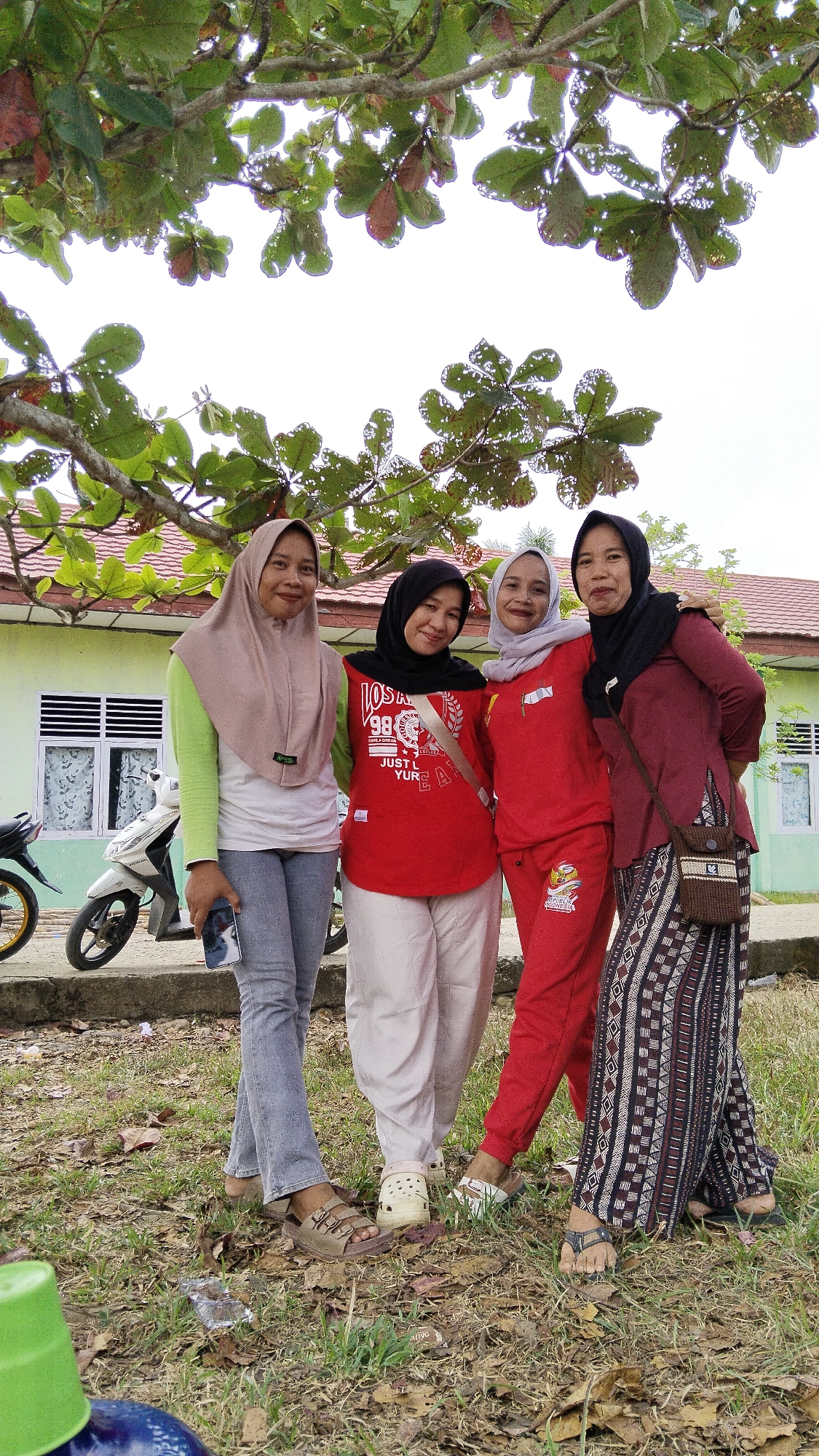 Four women are posing for a photograph outdoors. They are standing on grass in front of a light green building with a red tiled roof. A tree with green leaves, some of which are turning red, is in the foreground, partially obscuring the sky. The woman on the far left is wearing a light brown hijab, a lime green long-sleeved shirt, and light gray jeans. She is holding a smartphone in her left hand. The second woman from the left is wearing a black hijab, a red t-shirt with graphics, and off-white pants. The third woman from the left is wearing a light gray hijab, a red long-sleeved shirt, and red pants. The woman on the far right is wearing a black hijab, a maroon long-sleeved shirt, and a patterned skirt. She has a small woven bag with brown and white accents slung over her shoulder. All four women are smiling and looking at the camera. To the left of the women, a white motorcycle is parked. The ground is covered in dry leaves and grass. The overall scene suggests a casual gathering or outing.
