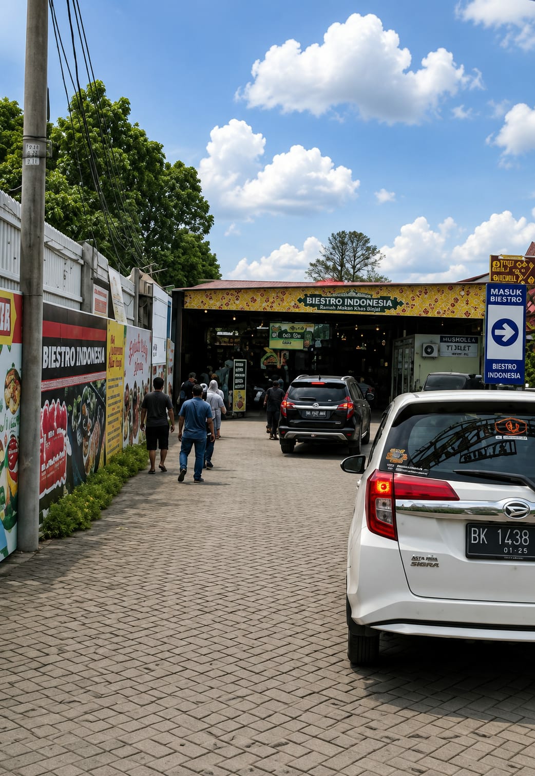 The image shows the exterior of a "Biestro Indonesia" establishment in the city of Binjai, Indonesia. The establishment appears to be a restaurant or food court, with banners and signage indicating food and drinks are available. Several people are walking towards and inside the building. There are also cars parked in front, suggesting a busy atmosphere. The architecture is open-air, with a yellow and red patterned awning extending over the entrance. The ground is paved with interlocking bricks. A blue directional sign with an arrow points towards "MASUK BIESTRO" (Enter Biestro). The sky is blue with scattered white clouds, and lush green trees are visible in the background. A white car with license plate "BK 1438" is parked in the foreground, facing away from the viewer.
