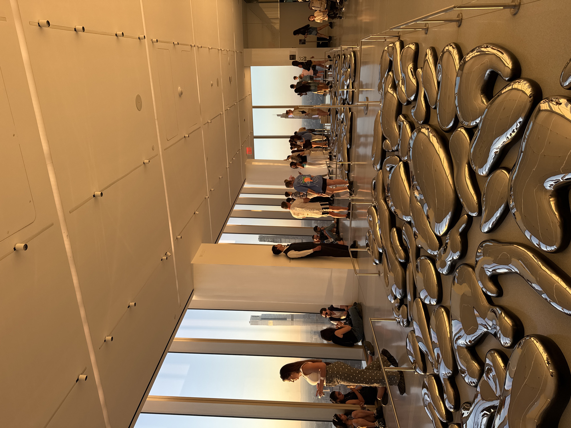 The image depicts the interior of a modern, high-rise building, likely an observation deck or an art exhibition space, with panoramic views of an urban landscape. The setting is New York City, indicated by the visible cityscape with tall buildings. The time of day appears to be late afternoon or early evening, with a warm, golden light from a setting sun casting a glow through the large, floor-to-ceiling windows.
Numerous people, casually dressed, are present in the space. Many are gathered near the windows, observing the city skyline. Others are interacting with or admiring a prominent art installation on the right side of the room. This installation comprises a large collection of irregularly shaped, highly reflective metallic objects arranged on a wall, which mirror the light, people, and the exterior view. Some individuals are seen using smartphones, likely taking photographs of the view or the art.
The interior features a high ceiling with recessed lighting, and light-colored walls. A white rectangular pillar stands near the windows, with the word "WEST" faintly visible, inscribed vertically on its surface. On the back of one person's t-shirt, the word "PASADENA" can be seen. The overall atmosphere seems relaxed and contemplative as visitors take in the expansive views and the art exhibit.