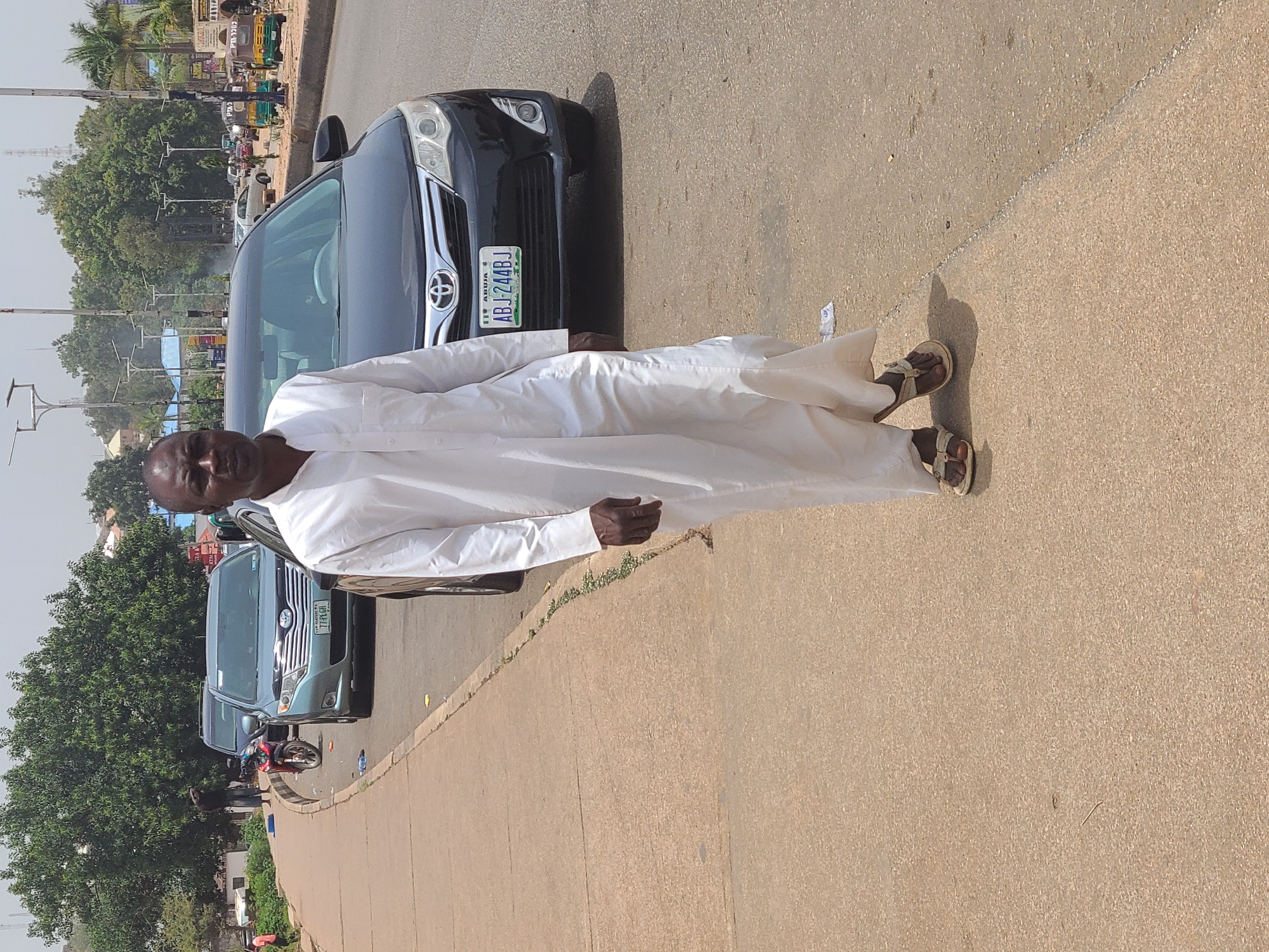A man dressed in a long, white garment and sandals stands on a city street. He is positioned adjacent to parked cars. The license plate on the nearest car is partially visible, showing "ABJ-244BJ" and indicating registration in Abuja, Nigeria. Other vehicles are parked further down the street. A line of trees is visible in the background. The location appears to be urban with a road, vehicles, and pedestrian activity. The location is likely in the city of Abuja, Nigeria.