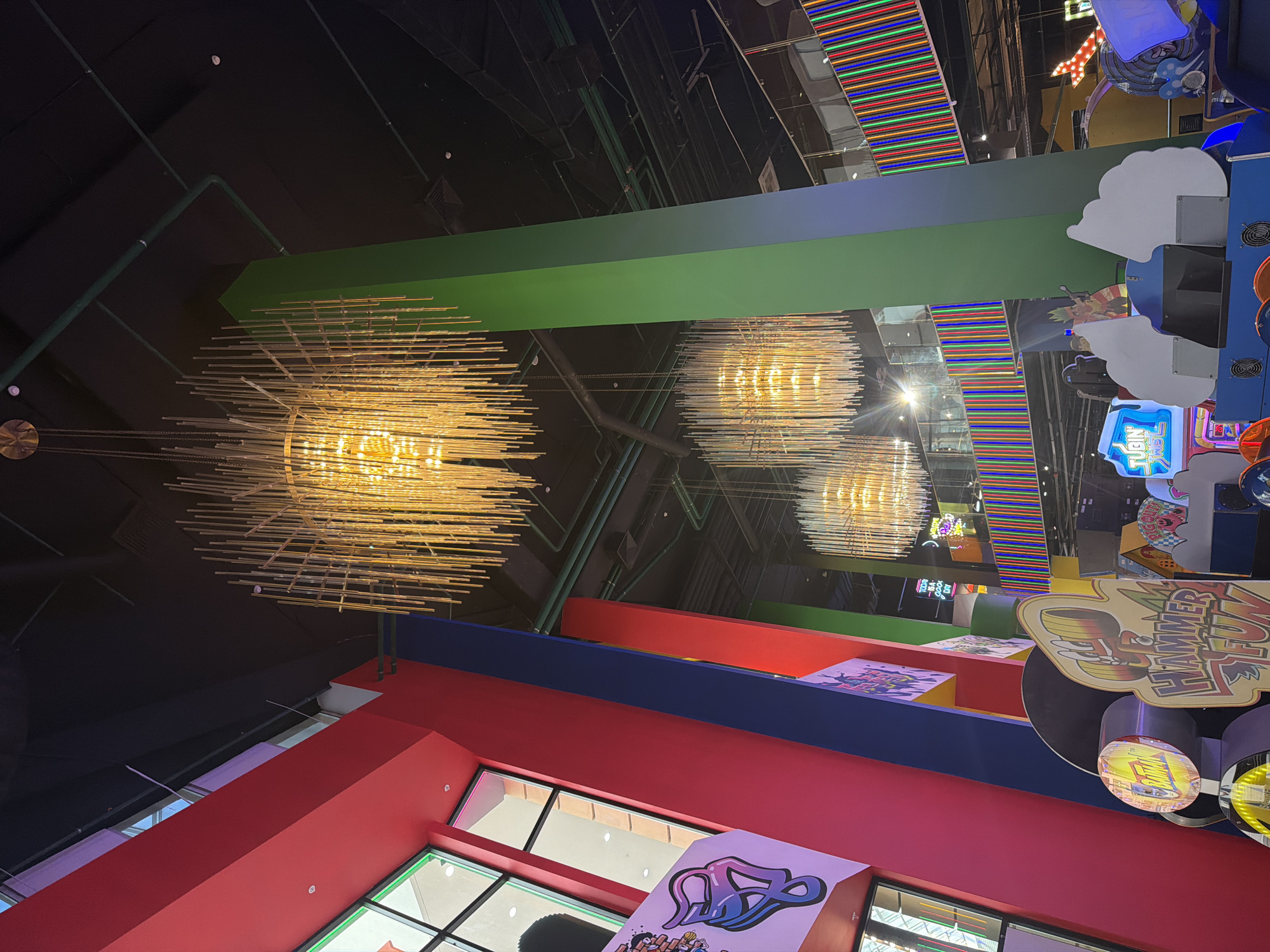 The image depicts an indoor arcade with a vibrant and colorful aesthetic. Dominating the foreground are several large, modern chandeliers with a multitude of thin, vertical rods that emit a warm, golden light. These chandeliers hang from a dark ceiling, which is crisscrossed with exposed green pipes and conduit.
Several large, brightly colored pillars are prominent in the scene. One pillar on the right is a gradient of green to blue. On the left, pillars are painted in bold red and blue hues. These pillars are adorned with graphic art, including a stylized purple and pink design on the left.
In the lower portion of the image, various arcade game facades are visible. One sign reads "HAMMER FUN" in large, playful lettering. Other games have illuminated signs with titles such as "TUBIN TWIST" and "BATMAN". The games themselves appear to be a mix of modern and classic designs.
In the background, multiple levels of the arcade are visible, separated by railings adorned with colorful LED lights in vertical stripes of red, green, blue, and yellow. The overall environment suggests a lively entertainment venue. No people are clearly visible in the image. The location is identified as Unknown, Unknown.
