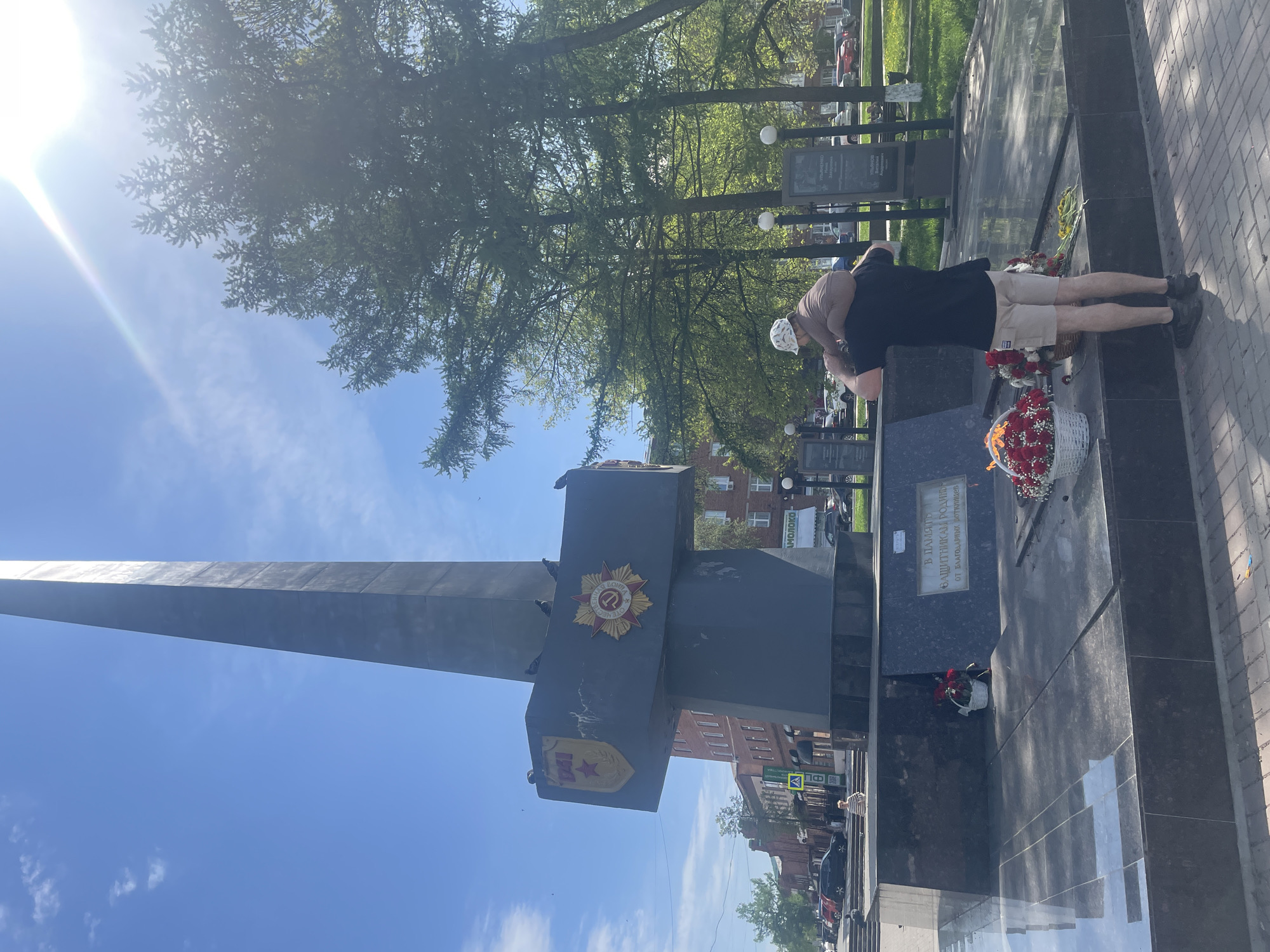 A memorial complex dedicated to the Victory in the Great Patriotic War is the primary focus of the image. The complex includes a prominent stele resembling a sword, adorned with Soviet emblems. The location is Votkinsk, Russia.
Two individuals stand at the base of the monument, near floral tributes. One person, in light-colored shorts and a dark shirt, appears to be interacting with the monument in a respectful manner.
The surroundings include trees, other memorial plaques, a paved area, and buildings. The sky is blue with some clouds and a bright sun.