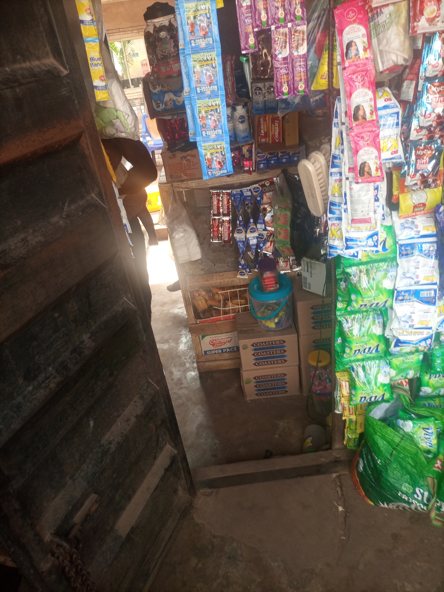 This is an indoor, medium shot of a small shop in Nkpor, Nigeria. The shop appears to be a convenience store or a small market stall, filled with various products. The scene is cluttered with merchandise displayed on shelves and hanging from the ceiling.
In the foreground, a large, dark wooden door or shutter is partially open, casting a shadow. Behind it, a person is visible, mostly obscured by the products, wearing dark pants and a light-colored shirt. They are standing in the shop, possibly attending to customers or organizing goods.
The shelves are packed with a variety of items, including packaged foods, drinks, toiletries, and cleaning supplies. Prominent brands and product names like "Blue Band," "Lucozade," "Glucose-D," and "Viva Plus" are visible on the packaging. Boxes labeled "Coasters" are stacked on a lower shelf. A blue bucket with a purple lid sits on top of some boxes.
The lighting suggests it is daytime, with sunlight entering from an opening in the back of the shop, creating bright spots and shadows. The overall impression is of a busy, functional retail space common in many Nigerian towns. The presence of various food items, beverages, and household necessities indicates the shop serves the daily needs of the local community. The rough, wooden structures and the density of products create a sense of a well-stocked, albeit modest, establishment.