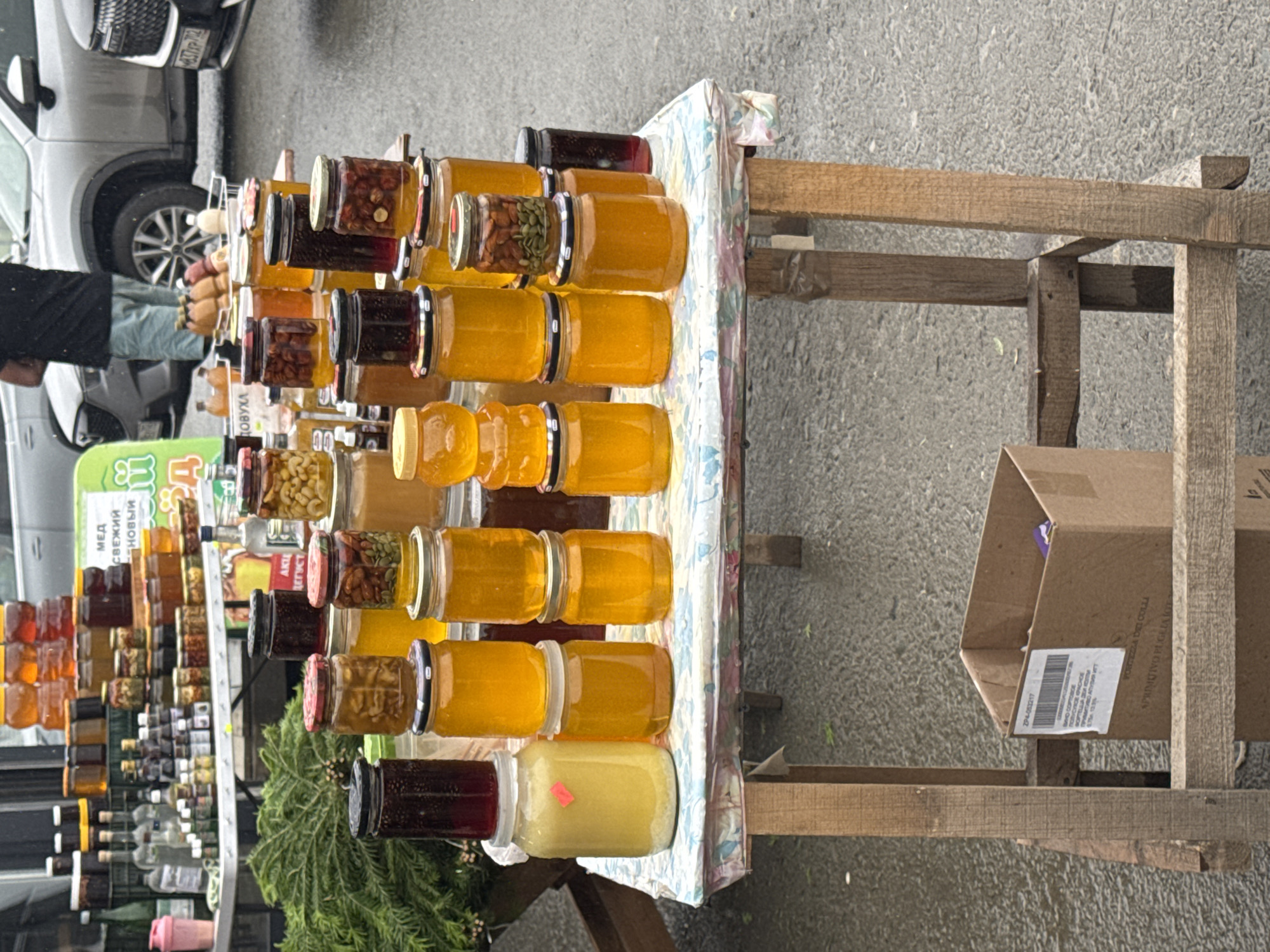 In Inzer, Russia, a market stall displays a variety of jars of honey and preserved goods. The items are arranged on a rustic wooden table covered with a patterned tablecloth. The jars contain honey of different shades of yellow and amber, as well as some with preserved fruits or nuts. Some jars are stacked on top of each other, creating vertical arrangements. In the background, a person is partially visible standing near a vehicle, and other market elements, such as a green banner with text and more bottles on a higher shelf, are present. The ground appears wet, suggesting recent rain. A cardboard box sits beneath the table.
