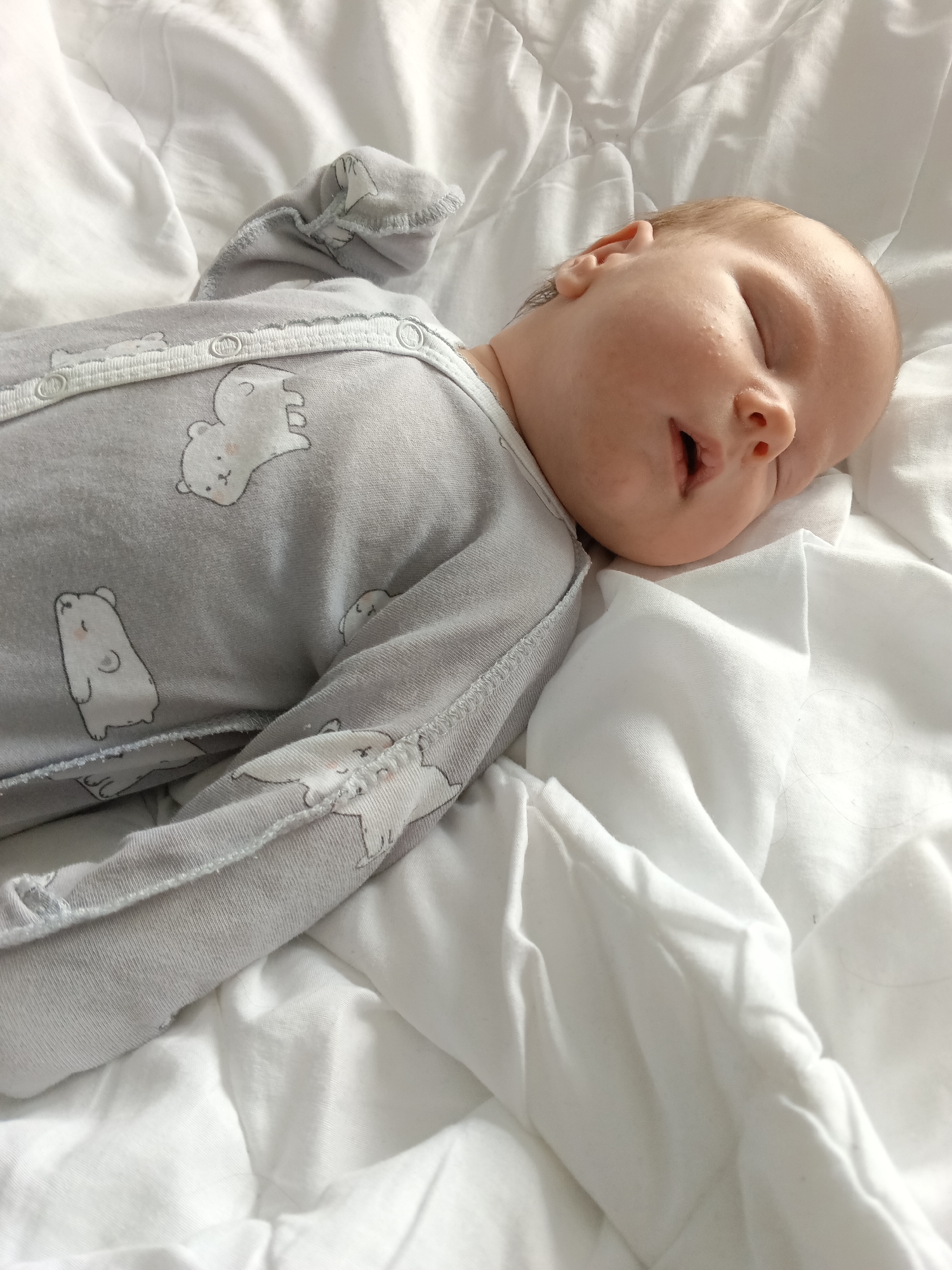 A baby is captured sleeping peacefully on a white bed. The infant lies on their back, turned slightly to their right side, with their head resting on soft, white bedding that appears somewhat wrinkled. Their eyes are closed, and their mouth is slightly agape. The baby has light, fine hair on their head. They are dressed in a light grey, long-sleeved onesie or sleepsuit adorned with a pattern of white cartoon bears. Snaps along the side of the garment are visible, securing it. The baby's skin shows a few small bumps or textures on the forehead and cheek. The overall atmosphere of the scene is calm and restful. The setting is indoors, likely a bedroom, in Andreyevo, Russia. No text is visible in the image.