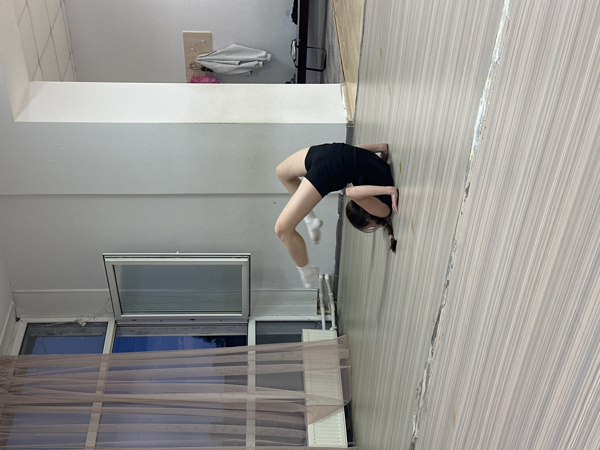 A girl is in a backbend pose in a room in Votkinsk, Russia. She is wearing black shorts and a black t-shirt with white socks. Her hands are resting on the floor. The room has light-colored walls and a window with curtains. There is a radiator beneath the window. The floor is made of light-colored wood.