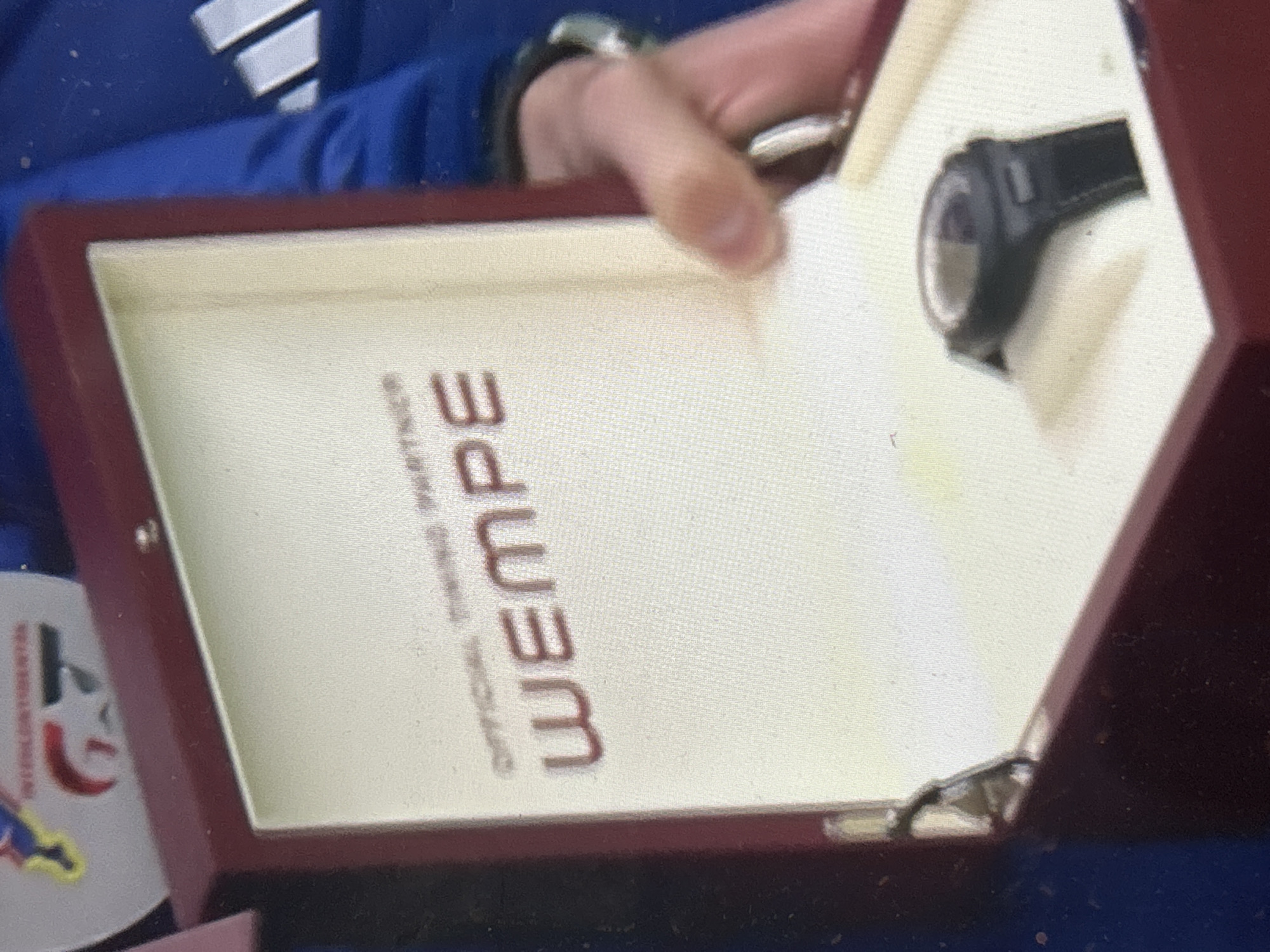 A partial view of a person in a blue jacket holds open a red wooden box containing a black wristwatch on a white cushion and the word "WEMPE" printed vertically in red on the interior lining. A second watch is visible on the person's wrist. The location is not discernible from the provided image.
