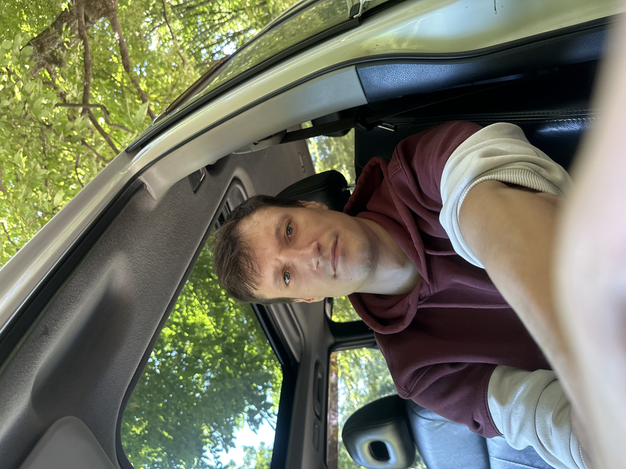 A man is sitting in the driver's seat of a car, looking at the camera. The car's sunroof is open, and sunlight streams through it, illuminating the interior. Trees and foliage are visible through the sunroof and the car's windows, suggesting an outdoor setting. The man is wearing a burgundy hoodie with light-colored sleeves. The interior of the car is visible, including the black leather seats and the car's headliner. The image is a selfie, taken from the man's perspective. There is no indication that this image was taken in Shatava, Ukraine.
