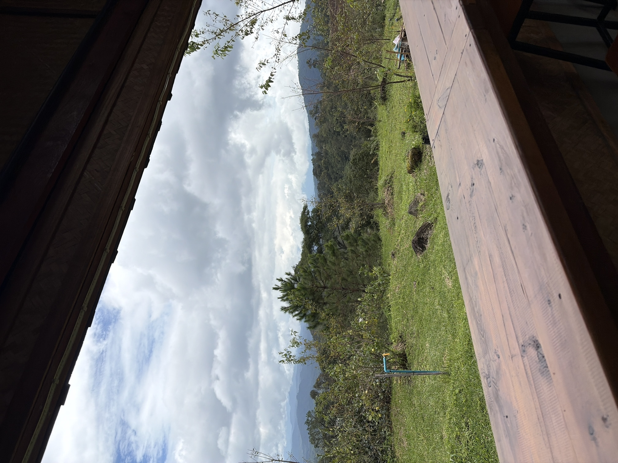 The image shows a view from a wooden balcony or deck overlooking a lush green landscape. In the foreground, a weathered wooden surface suggests a table or railing. Beyond this, a grassy slope leads to a variety of trees and vegetation. A prominent pine tree stands to the right of center, with a blue pipe and faucet visible in the grass to the left. In the distance, rolling hills covered in dense forest stretch towards a cloudy, overcast sky. The overall impression is of a natural, rural setting, possibly in a mountainous region. The location is identified as Ban Khun Wang, Thailand, suggesting a cool, high-altitude environment known for its agriculture and natural beauty. There are no people or animals visible in the frame.