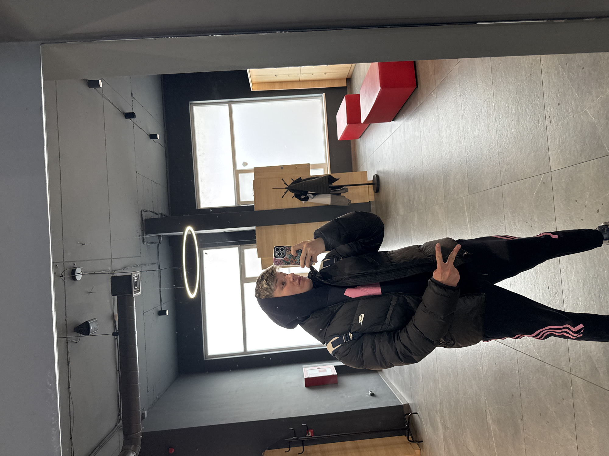 The image displays a full-length mirror reflection of a young male adult, centrally positioned, engaged in taking a selfie within an indoor environment in Plovdiv, Bulgaria. The male is dressed in a black hooded sweatshirt with the hood up, a black puffer jacket bearing a white Nike swoosh on the left chest, and black athletic trousers with three parallel pink stripes down each outer leg. He holds a smartphone, equipped with a multi-colored, patterned case, in his right hand, angled to capture his reflection. His left hand is raised, forming a "peace sign" gesture. The background depicts a modern, industrial-style interior featuring dark grey walls and light grey tiled flooring. The ceiling is grey with visible metallic ventilation ductwork, multiple black track lights, and a prominent white circular LED light fixture. To the left of the subject, a black metal coat rack holds dark and white garments, and a red wall-mounted fire extinguisher cabinet is present. Further into the background, two rectangular red upholstered benches are positioned parallel to each other, in front of a wall with large frosted windows. Light wooden lockers are partially visible on the far right side of the frame.