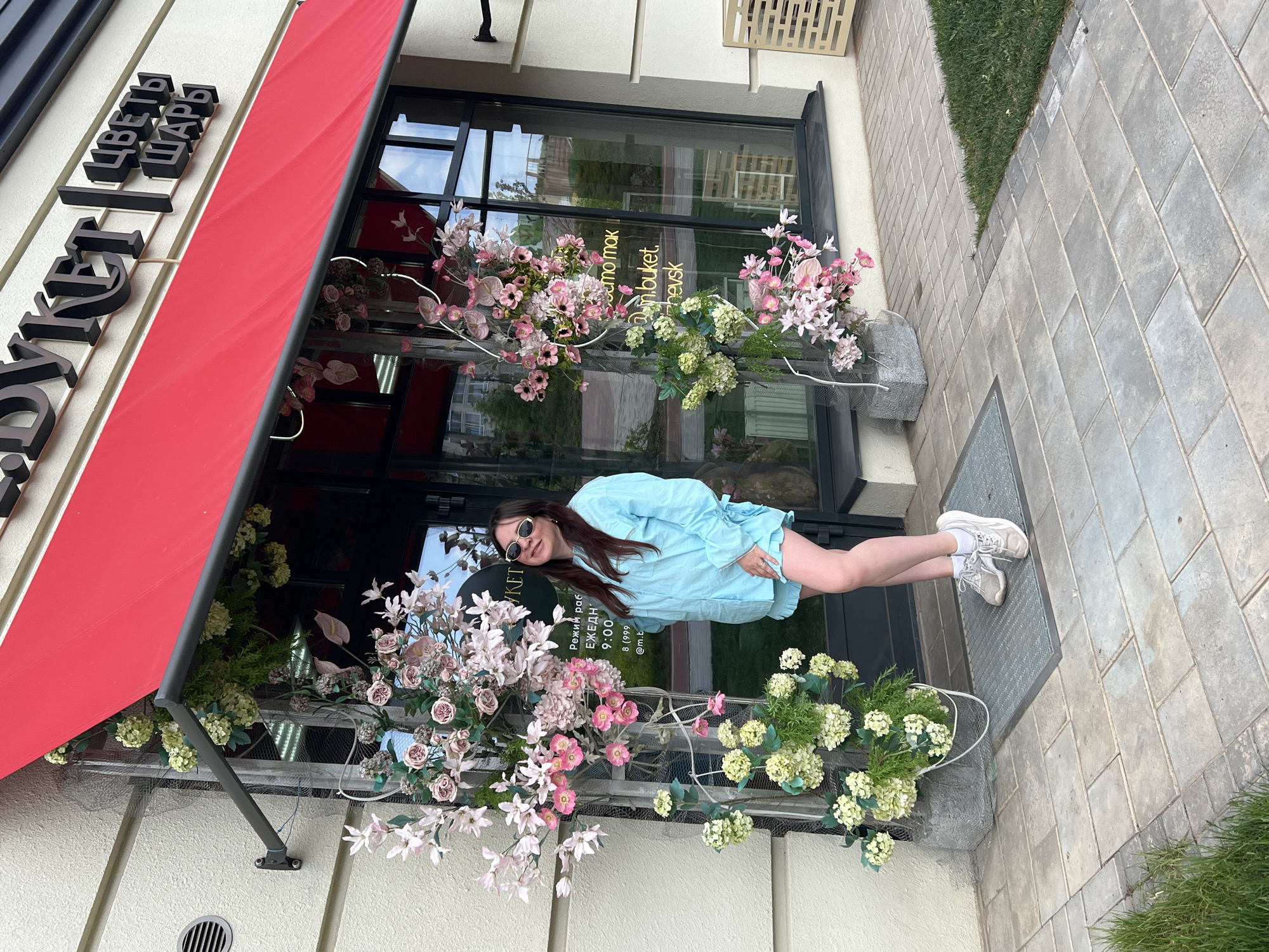 A slightly rotated, medium shot depicts a woman standing in front of a flower shop entrance in Pervomaysky, Russia. The shop has a red awning and is decorated with pink and white flowers. The woman wears a light blue dress and white sneakers. She is smiling and wearing sunglasses. The facade of the store is painted in bright colors. A sign in Russian above the awning reads "Buket" and "Tsvety Shary" (which means "Bouquet" and "Flowers Balloons"). The sidewalk in front of the shop is paved with gray stones.