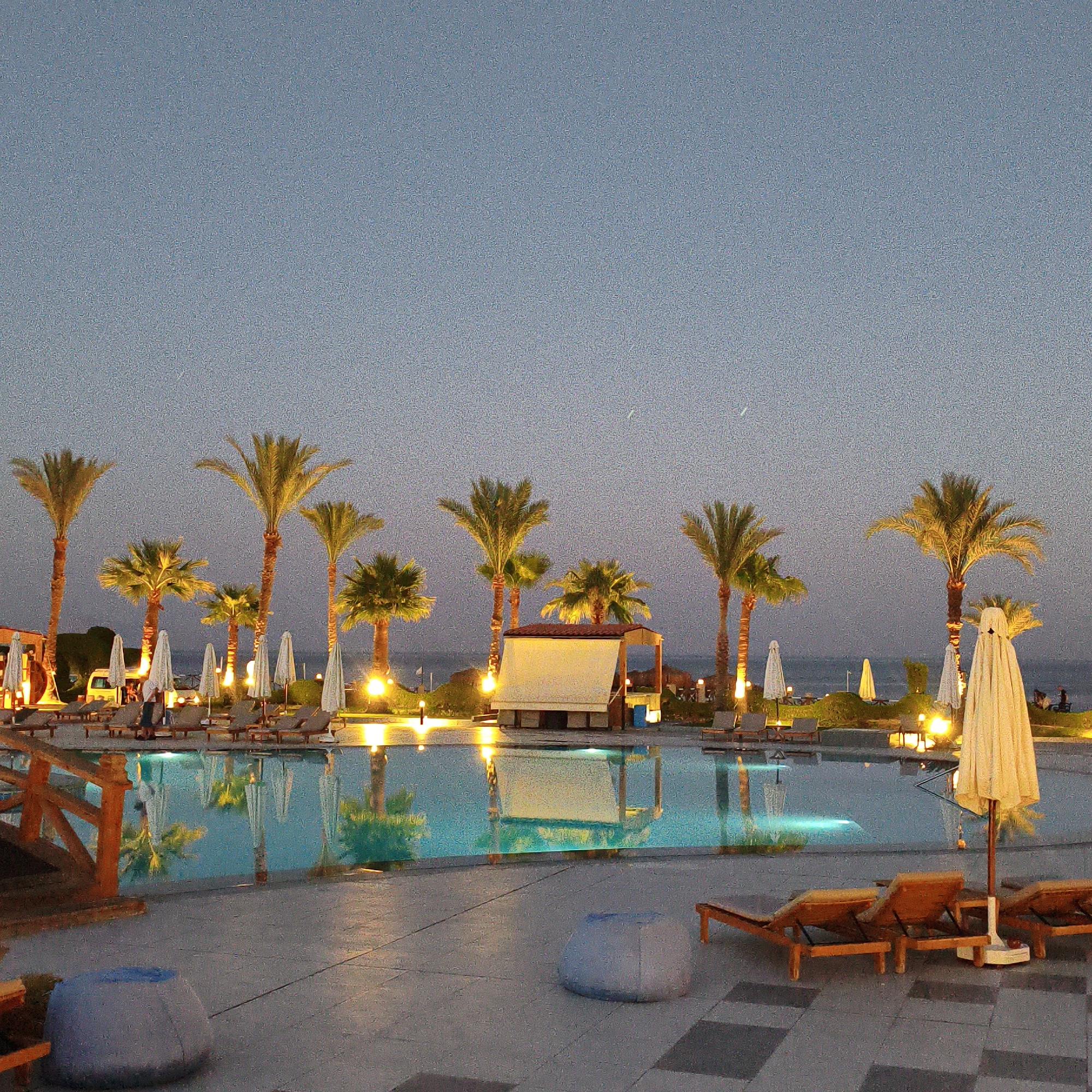 The image depicts a resort area at dusk. In the foreground, there is a swimming pool with lounge chairs and umbrellas surrounding it. The pool water reflects the lights and palm trees. Several palm trees are scattered throughout the scene, illuminated by warm lighting. In the background, the ocean is visible, and a small building with a thatched roof is situated near the pool. The sky is a dusky blue, with some faint celestial bodies visible. There are a few people in the distance, appearing to be walking or standing near the pool. The overall atmosphere is serene and tropical.