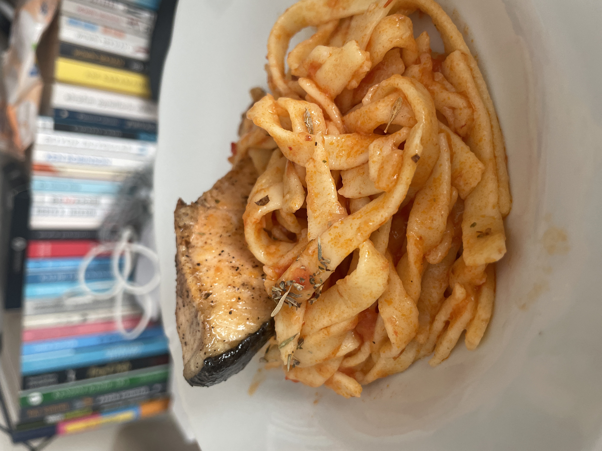 A close-up, eye-level, slightly angled shot captures a white plate filled with pasta and a piece of salmon. The pasta, a wide, flat noodle shape, is coated in a reddish sauce and sprinkled with herbs. A piece of salmon, seared with a dark, crispy skin and a flaky, orange interior, is positioned on top of the pasta. The background is out of focus, showing a bookshelf filled with colorful books. The scene suggests a meal, possibly in a home or restaurant setting. There is no identifiable location in the city of Başakşehir, Turkey, depicted in the image.