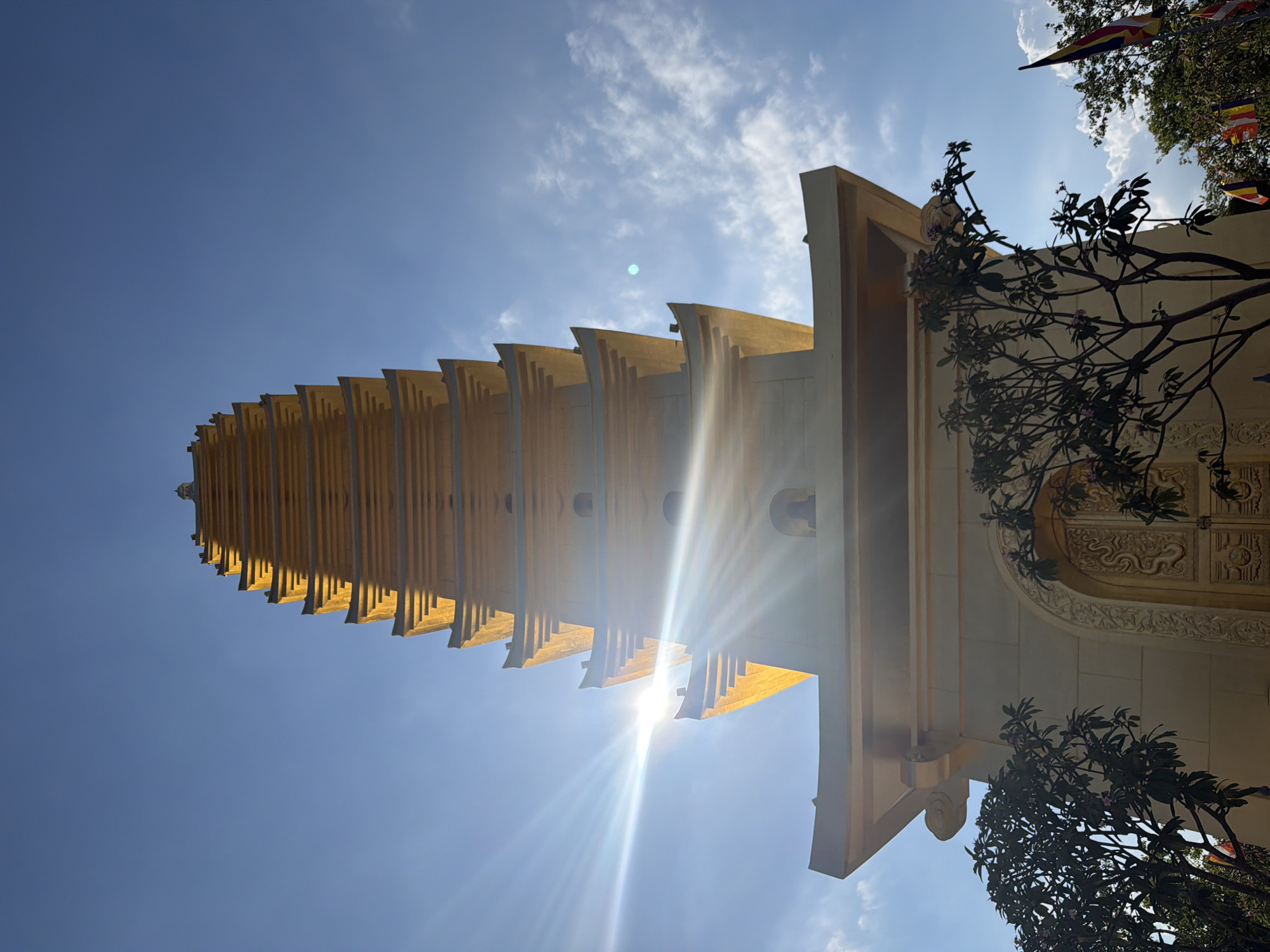 A low-angle, upward-facing shot of the gold-colored Long Son Pagoda in Nam Nha Trang Ward, Vietnam, against a bright blue sky with scattered clouds. The pagoda, which takes up the middle and left side of the shot, extends into the sky with multiple stacked tiers. Sun glares brightly from behind the pagoda. To the right, part of the building's architecture is visible, along with trees. At the top right of the frame, there are Buddhist flags.