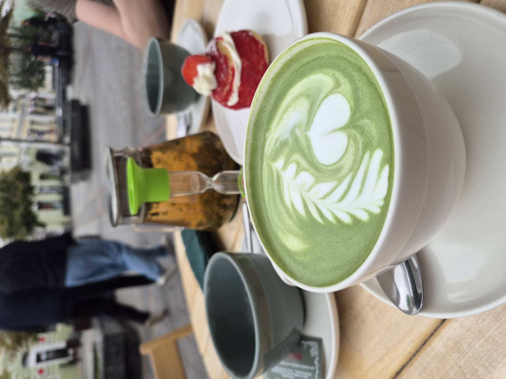 A close-up shot features a white mug filled with vibrant green matcha latte, adorned with intricate foam art resembling a fern. The mug rests on a white saucer, accompanied by a silver spoon. To the left, a grey ceramic bowl sits on a small white plate with a green booklet. In the background, a glass pitcher holds amber-colored liquid with dried herbs, and a small hourglass with a green top stands beside it. A red pastry topped with whipped cream and a strawberry slice is visible on a white plate to the right. The scene is set on a wooden table, with blurred figures and buildings in the distance suggesting an outdoor cafe setting in Vladivostok, Russia.
