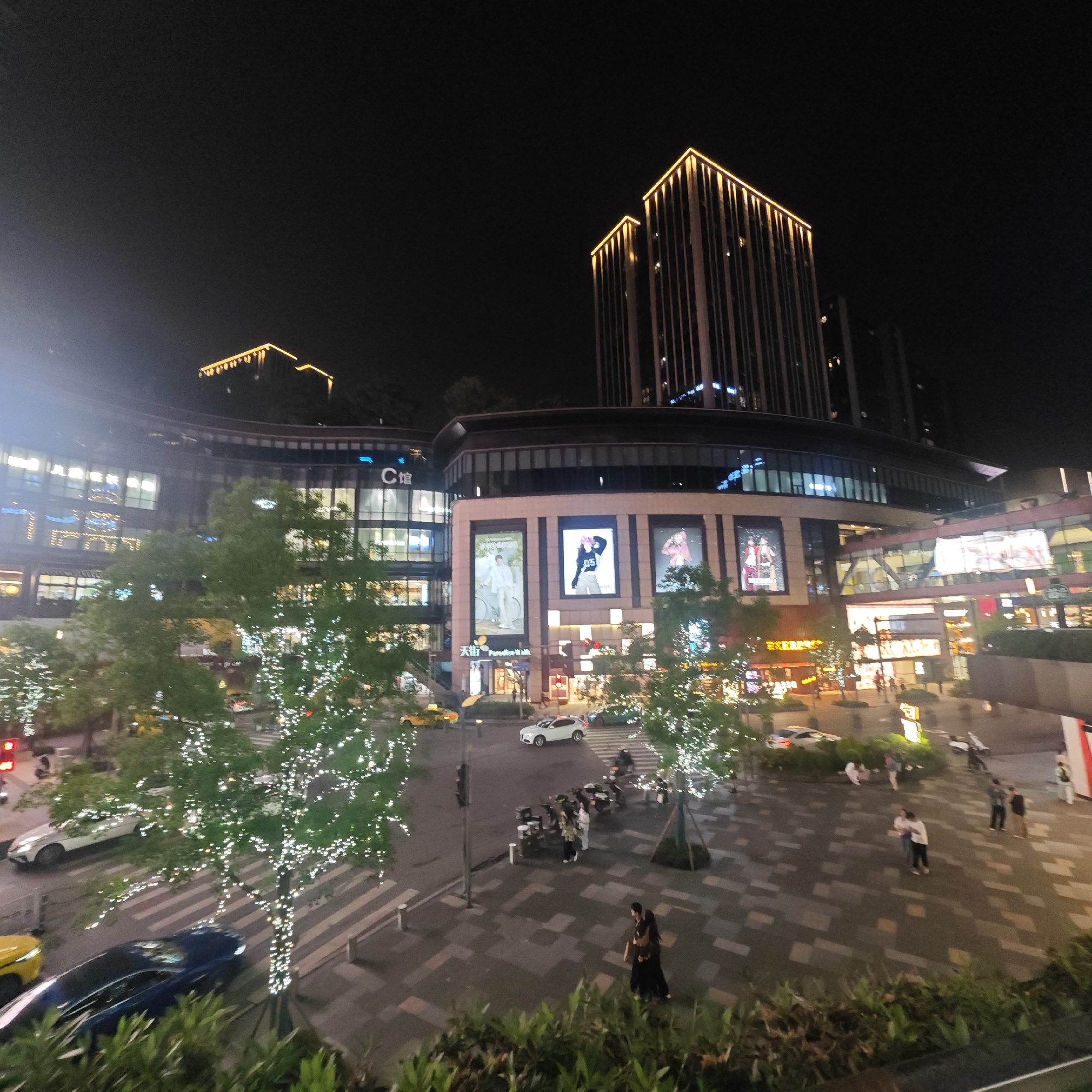 This media file presents a vibrant night scene of a modern commercial district, likely located in Yuzhong District, China. The setting is dominated by contemporary multi-story buildings, some featuring striking illuminated golden lines along their upper structures, creating a sleek, urban aesthetic. One prominent building clearly displays the characters "C 馆" (C Guǎn, meaning C Block/Hall) and features large, bright advertising screens showcasing models. Below these screens, the text "天街" (Tiān Jiē) and "Paradise Walk" is visible, indicating a shopping street or complex. A pedestrian walkway or bridge connects parts of the buildings.
The foreground and midground feature a street bustling with traffic, including a yellow taxi, a white SUV, and a blue sedan. Pedestrians are scattered across the paved areas and near crosswalks. The scene is adorned with numerous trees, intricately wrapped with strings of bright white lights, contributing to a festive and lively ambiance. Individuals and small groups of people are seen walking, standing, and interacting on the wide patterned pavement. The entire area is brightly illuminated by artificial lights emanating from the buildings, street lamps, and the decorative tree lights, starkly contrasting with the dark night sky. The image conveys a sense of a modern, active urban center during the evening.
