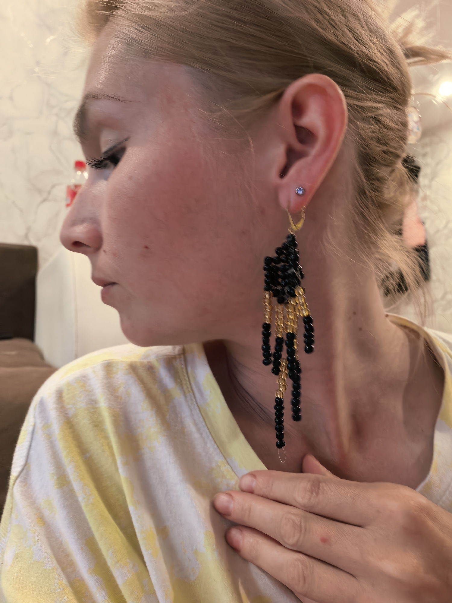 A woman with light-colored hair, styled in a ponytail, is shown in profile, facing left. She is wearing a yellow and white tie-dye t-shirt and a long, elaborate earring composed of black and gold beads hanging from her right ear. Her right hand is placed on her chest. The background is blurred, showing what appears to be an indoor setting with wallpaper and furniture. There are no identifiable landmarks that would suggest a location in Sterlitamak, Russia.