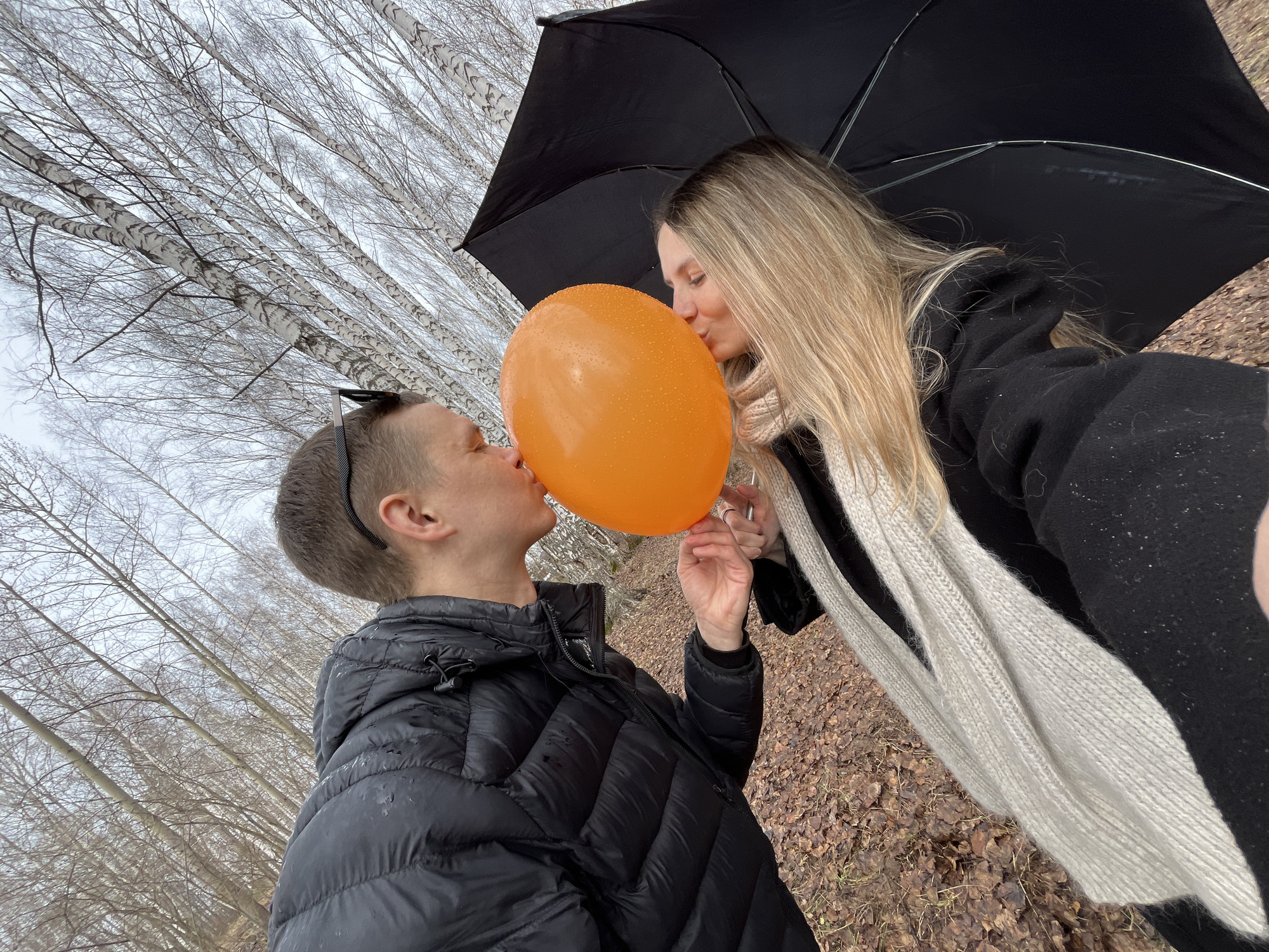 In the city of Chaikovsky, Russia, an outdoor photograph captures two individuals kissing an orange balloon held between them. The person on the left, with short brown hair, is wearing a black puffy jacket and sunglasses atop their head. The person on the right, with long blonde hair and a cream-colored scarf, is wearing a black coat and holding a black umbrella over them. The ground is covered in brown leaves. The background features a dense forest of birch trees with white bark and bare branches against a cloudy sky. The balloon has water droplets on its surface.