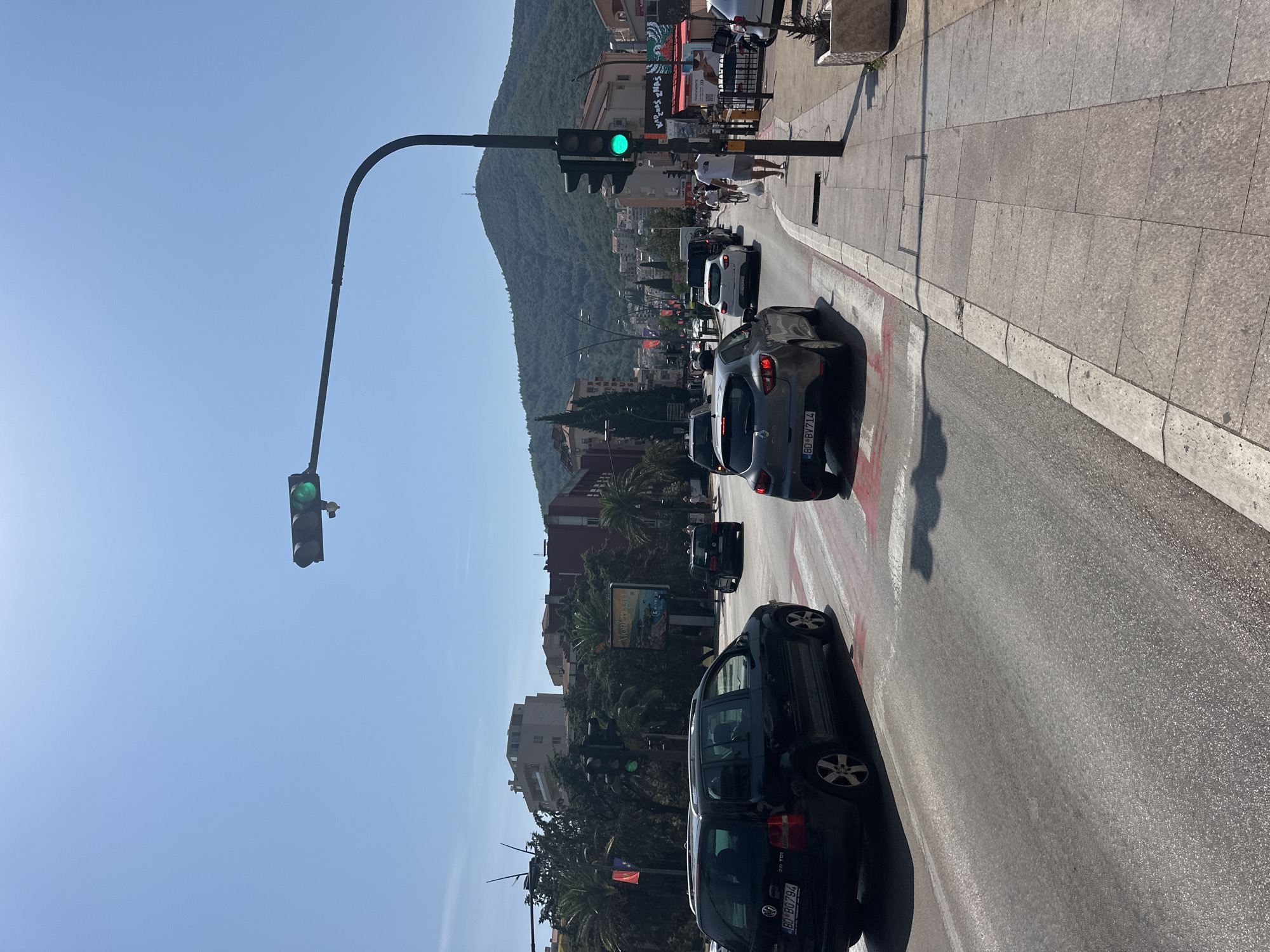 In Budva, Montenegro, on a sunny day with a clear blue sky, a traffic intersection is depicted. A traffic light overhead displays a green signal. Cars are navigating the road, including a black Volkswagen station wagon in the foreground, a grey Renault hatchback, and several other vehicles further back. Pedestrians are visible on the sidewalk, and a man in a white t-shirt walks with a shopping bag. Buildings line the street, with lush green trees and palm trees interspersed. In the background, a large, tree-covered mountain dominates the landscape. A billboard with the word "Montenegro" is also visible. The scene suggests a busy urban environment with ongoing traffic flow.