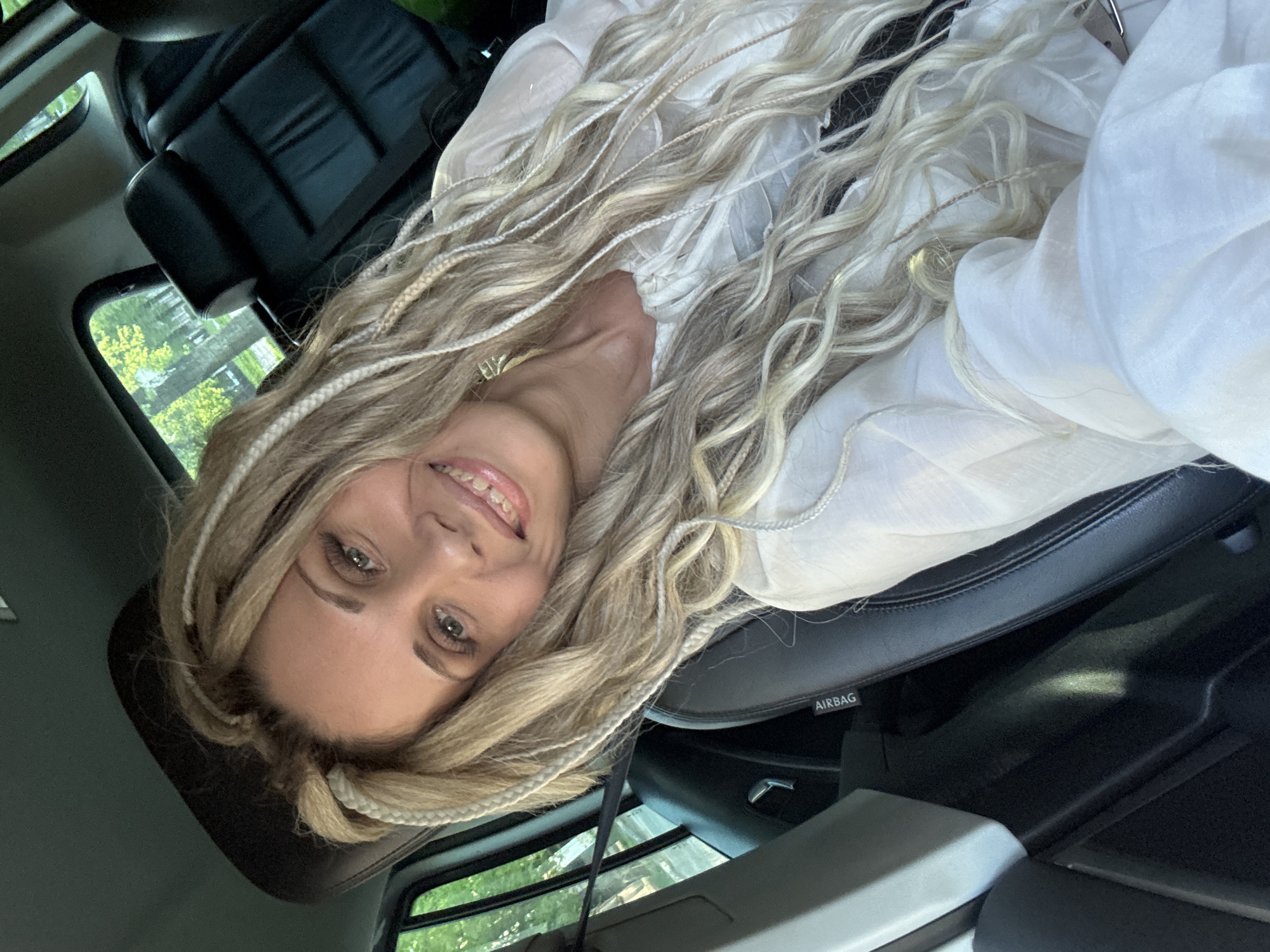 A woman with blonde braided hair is sitting in the driver's seat of a car. She is wearing a white blouse and smiling at the camera. The interior of the car is black leather. The environment appears to be outdoors with trees visible through the car window. The location, Rusanovo, Russia, is provided context, but not directly visible in the image.