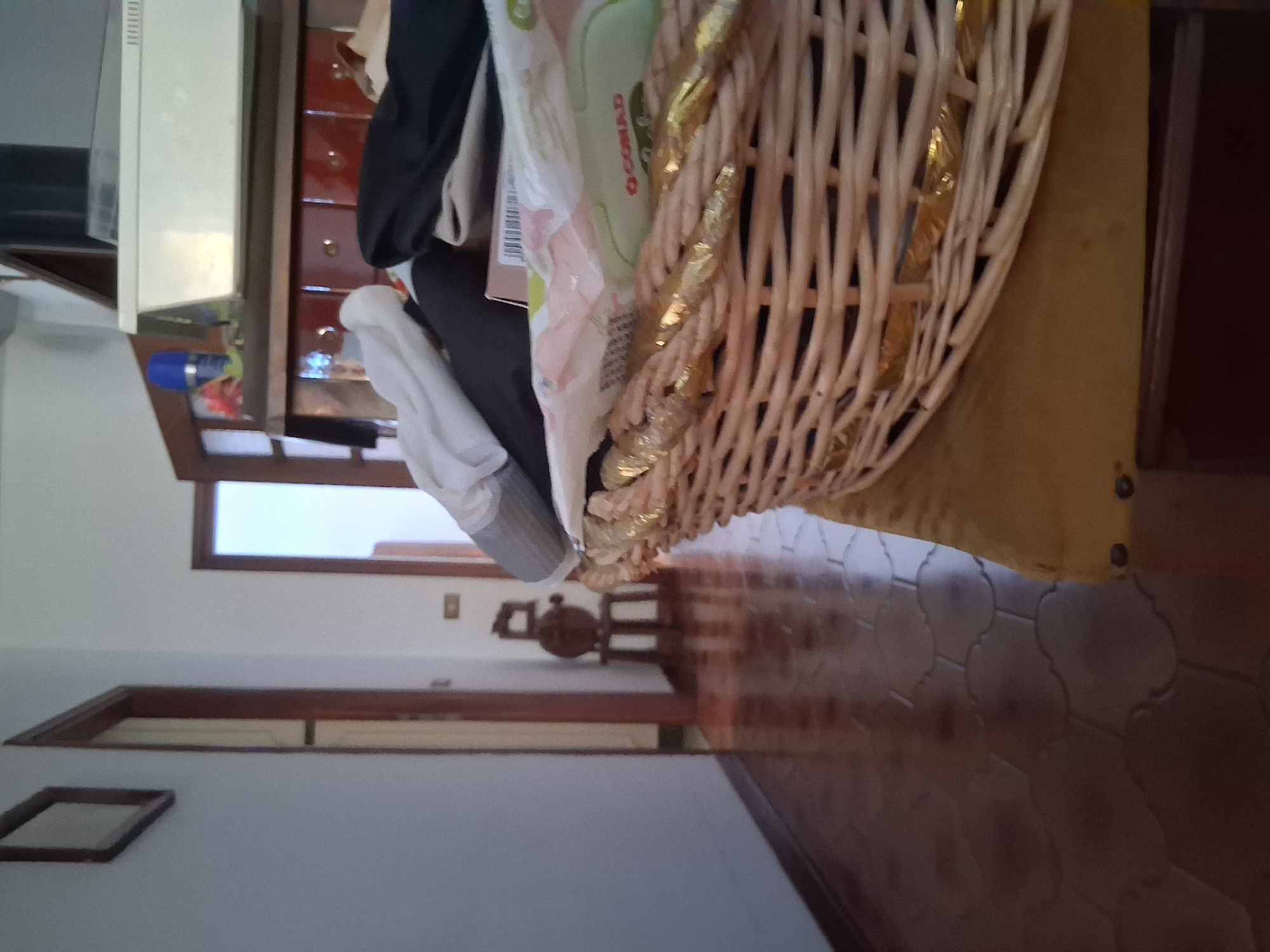The image shows an interior scene, likely a home in Colorno, Italy. A woven basket filled with various items, including what appears to be clothing and packaging, is prominently displayed. The basket has a decorative golden ribbon woven through it. To the left of the basket, a wooden cabinet with drawers is visible. A white appliance, possibly a refrigerator or washing machine, is partially visible on the far left. In the background, a hallway leads to a door, and there's a glimpse of a tiled floor. A small framed object is on the wall to the left, and a light switch is visible in the distance. The overall impression is of a domestic space with items stored in a basket. There is no discernible activity or event taking place; it appears to be a still life composition. The lighting suggests it is daytime, but no specific time of day or weather conditions can be determined. Text is visible on one of the items in the basket, appearing as "CONAD" printed on a green and white package.