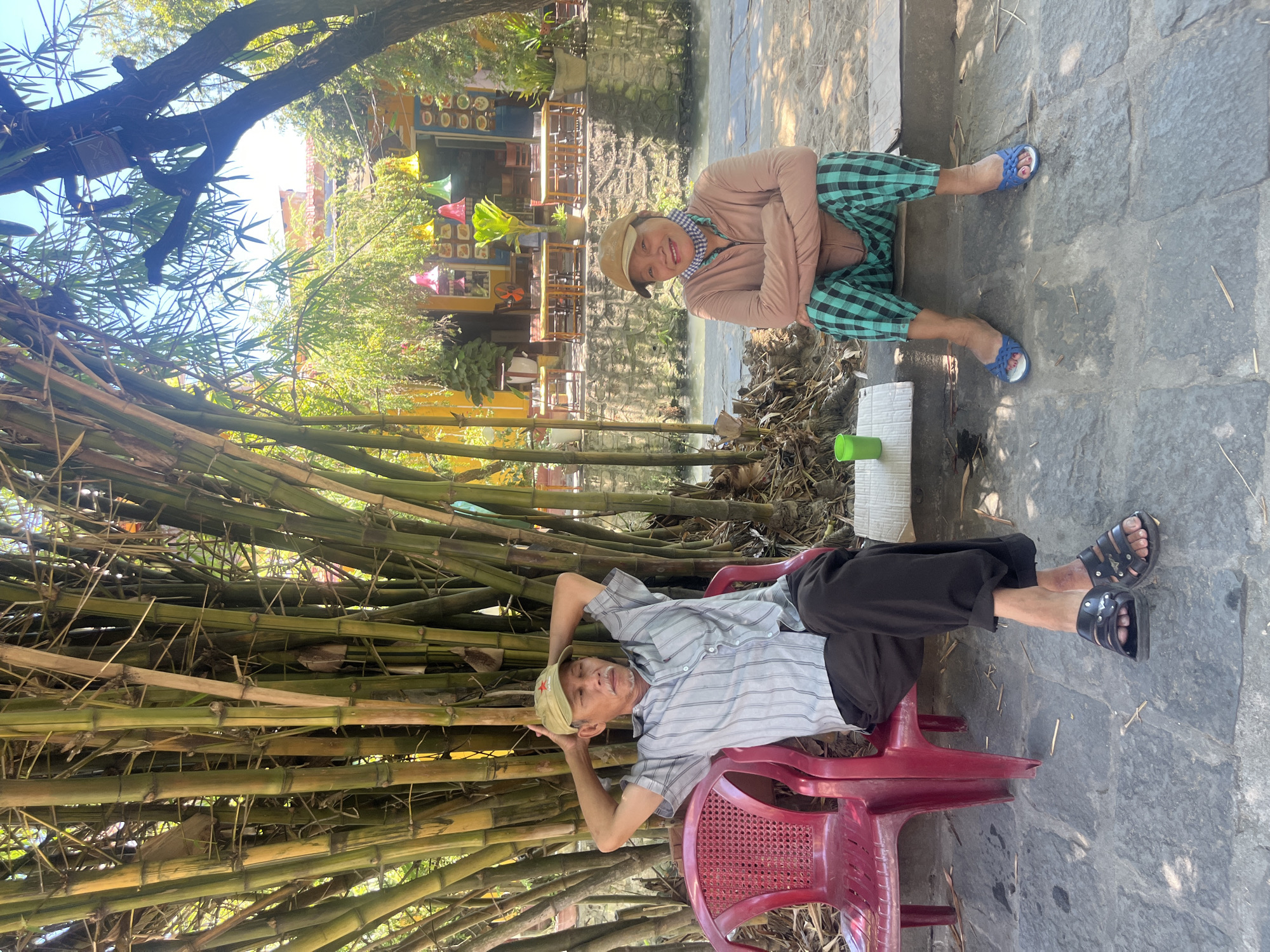Two individuals are resting outdoors on a sunny day in what appears to be Hội An Ward, Vietnam, by a body of water. The image is taken from a slightly elevated perspective, looking towards a canal or small river.
On the right side of the frame, an older woman is seated on a stone pavement, leaning back slightly and smiling towards the camera. She wears a long-sleeved light brown top, checkered green and black pants, blue sandals, and a light-colored cap with a patterned scarf around her neck. A green plastic cup rests on a white block beside her.
On the left, an older man is lounging comfortably in a red plastic chair, with his hands clasped behind his head. He wears a light-colored striped shirt, dark pants, and black sandals. A light-colored cap with a red star emblem sits on his head, and he has a mustache.
Behind the man, a dense grove of bamboo stalks creates a natural backdrop, providing shade. A narrow waterway separates the foreground from the background. Across the water, a building with yellow walls and multiple windows is visible. Colorful decorative lanterns (red, green, yellow) hang outside this building, which appears to be a restaurant or shop with outdoor seating. The ground is paved with large, irregularly shaped stones. Bright sunlight indicates it is daytime, and both individuals appear relaxed and at ease. No discernible text is visible in the image.