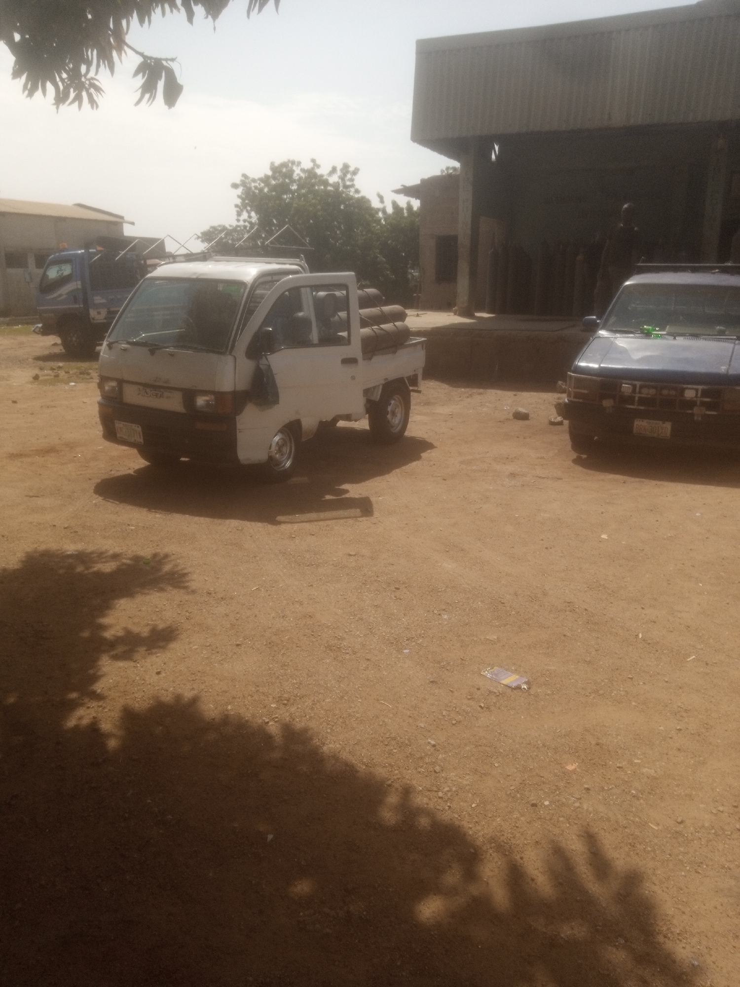 The image depicts an outdoor scene in Television, Nigeria, likely in a commercial or industrial area. Two vehicles are prominently featured. In the foreground and slightly to the left is a white minitruck, identified by a "Hijet" badge on its front. The driver's side door is open, and the truck bed contains several cylindrical objects, possibly gas cylinders, covered with a tarp. To the right of the minitruck, and further back, is a dark-colored pickup truck. In the background, a large, corrugated metal building with an open-front structure is visible, with numerous tall, cylindrical objects lined up inside, suggestive of a storage facility for gas cylinders. The ground is covered in dirt and sand. Overhead, tree branches with leaves cast shadows on the ground. The sky is bright and hazy, indicating a sunny day.