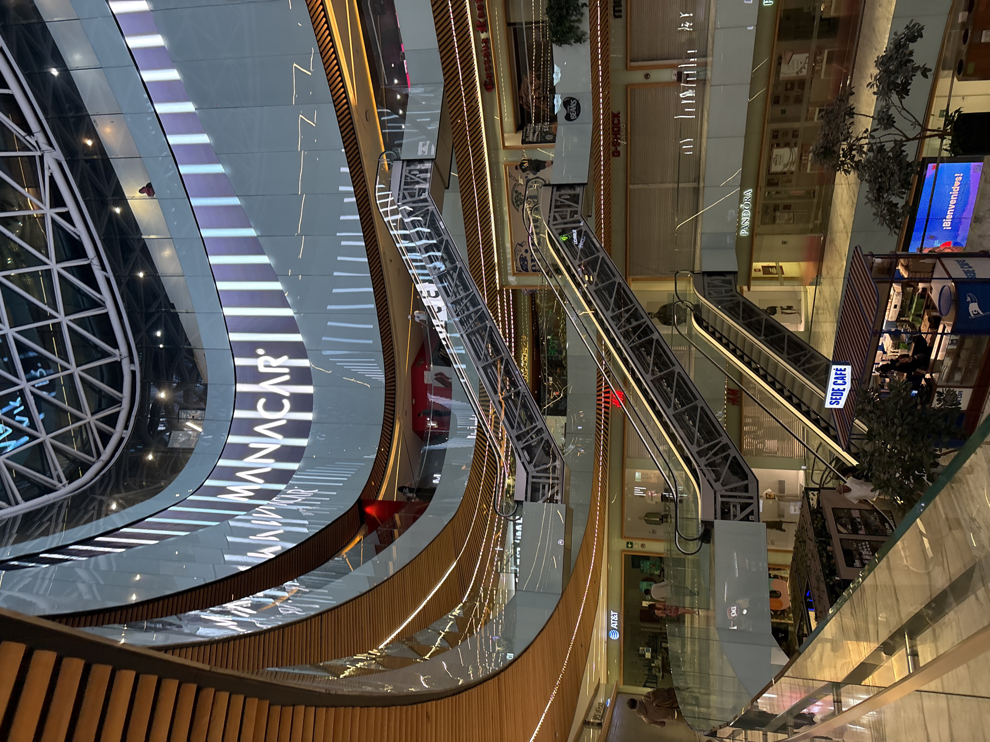The image provides an aerial view of a vibrant, multi-level shopping mall interior in Mexico City, Mexico, likely during the evening or night given the extensive artificial lighting. The scene is dominated by a grand, curving architectural design featuring numerous escalators crisscrossing a central void. Warm-toned wooden railings and glass barriers, illuminated by integrated light strips, guide visitors along the curved walkways and around the open space.
Digital display panels wrap around the upper levels, one prominently showcasing the text "MANACAR." Storefronts are visible on the various floors, some appearing closed with roll-down grates, while others display their wares. Identifiable brands include PANDORA, AT&T, G-SHOCK, CASIO, and BEANS. A small cafe, "SEDE CAFÉ," is active on a lower level, with patrons seated at tables. Another digital sign displays the Spanish greeting "¡Bienvenidos!" (Welcome!).
Several people are scattered throughout the mall, some walking along the corridors or riding the escalators, while others are dining at the cafe. Potted plants or small trees add greenery to the modern, brightly lit environment. The composition highlights the mall's expansive and sophisticated design.
