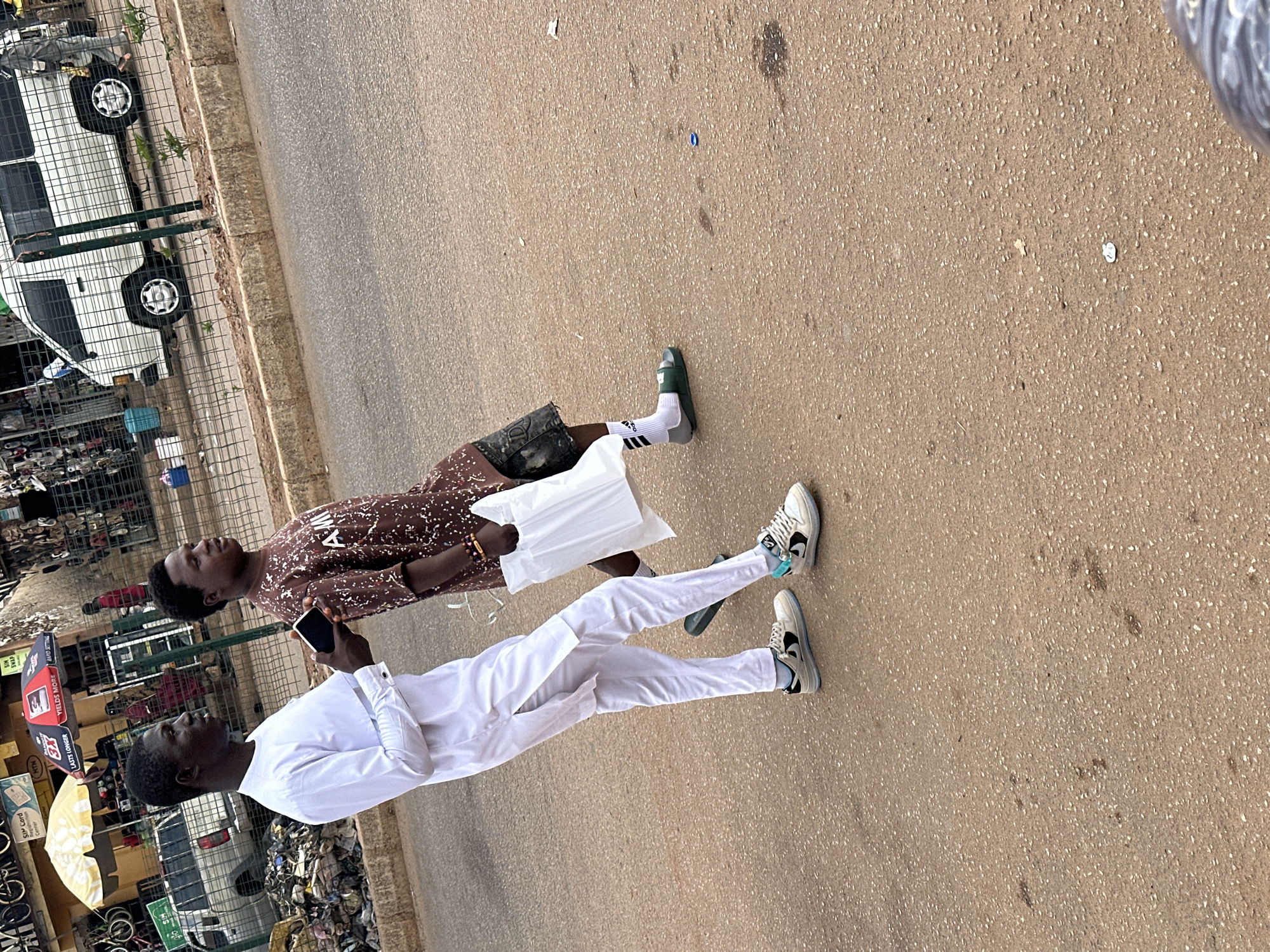 Two young men are walking on a paved surface. The man in front wears a white long-sleeved shirt and white pants. The man behind him wears a brown patterned shirt and dark shorts. They are walking on a street in what appears to be Bukuru, Nigeria, based on the presence of roadside kiosks. One man holds a phone. In the background are vehicles parked along the side of the road and commercial kiosks.