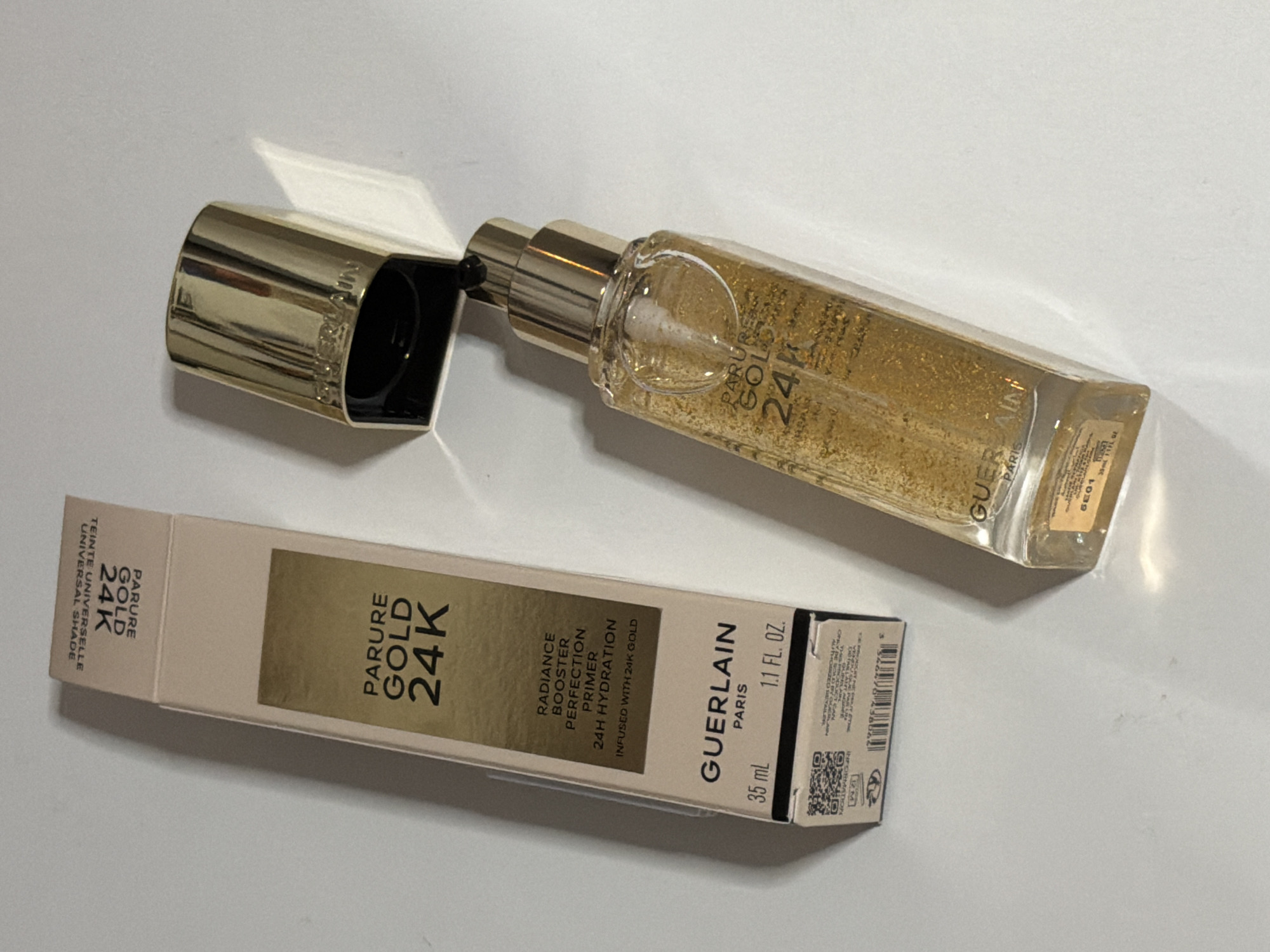 A gold-colored Guerlain Parure Gold 24K primer bottle and its packaging are displayed on a white surface. The bottle contains a clear liquid with gold flecks. The cap of the bottle is off and placed next to it. The packaging is a light beige box with gold lettering. The scene is illuminated by artificial light, casting shadows. There are no people or animals present. The location of this image is not specified in the provided information.