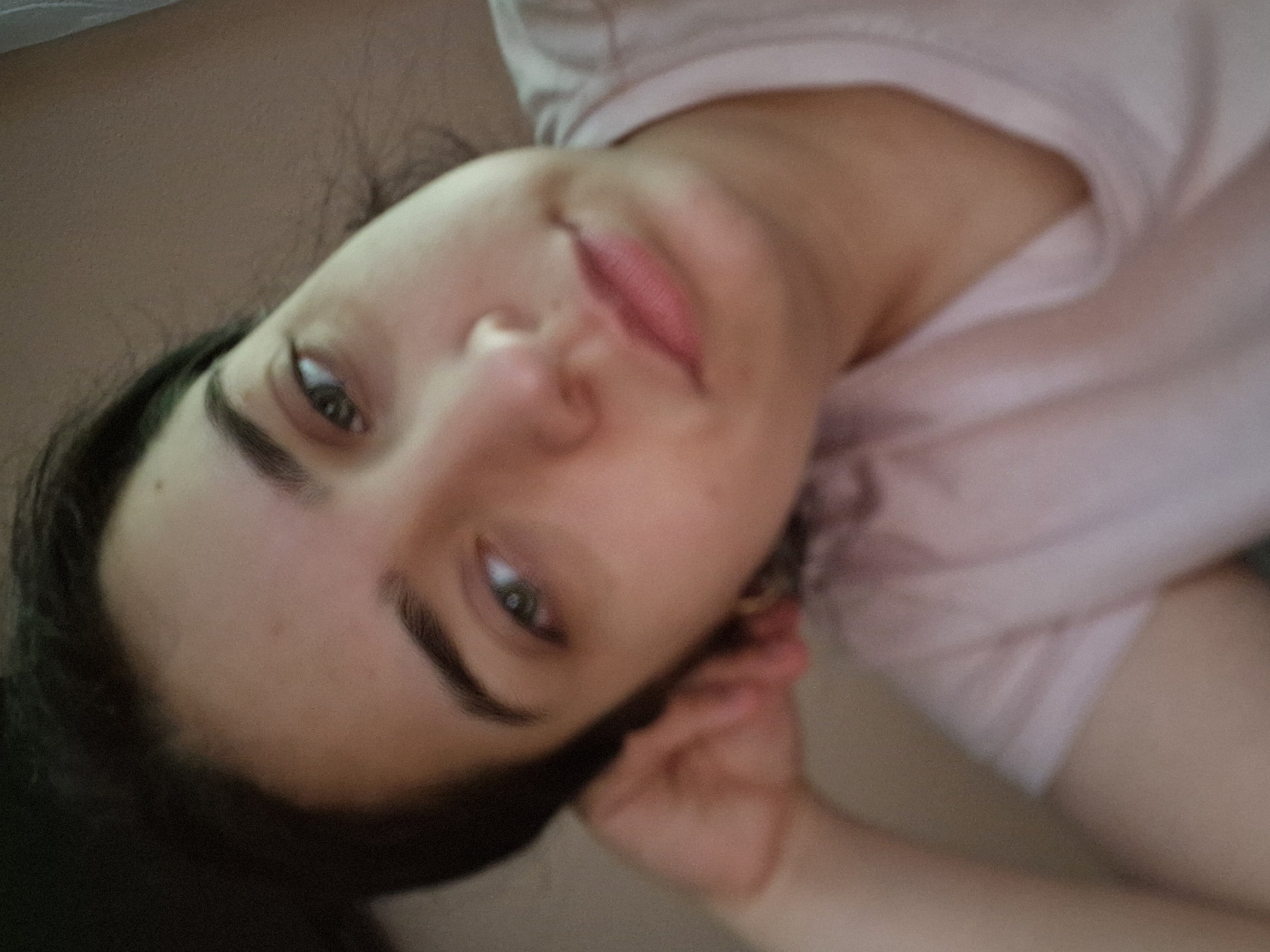 A close-up, slightly angled view shows a woman with fair skin, dark hair, and dark eyebrows. She has green eyes and pink lips. She is wearing a light pink t-shirt. Her right hand is near her ear. The background is a neutral, beige color. The location is unknown.