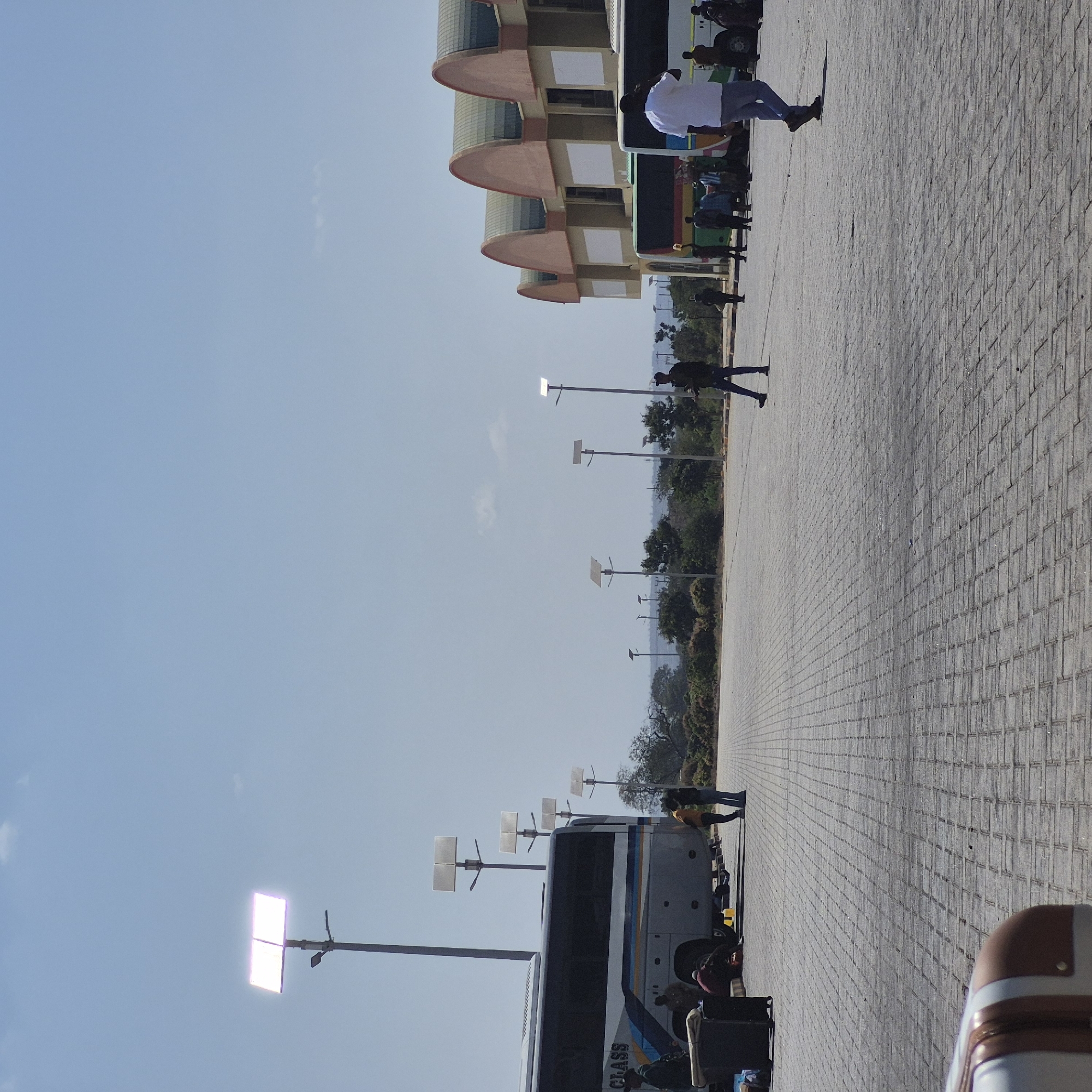 In Dodoma, Tanzania, an outdoor scene unfolds at what appears to be a bus terminal or waiting area. The ground is paved with interlocking grey bricks. In the background, a modern building with arched roof sections, painted in shades of orange and cream with white panels, is visible. Several buses are parked, including a grey and blue bus with "CLASS" written on its side. Solar-powered streetlights with square panels are scattered throughout the area, with some illuminated. Several people are present, some standing and conversing, others moving about the scene. One individual in a white shirt and blue pants walks away from the camera towards a bus, while another in a dark shirt and blue jeans walks towards the left of the frame. Luggage, including a suitcase, is present on the ground near the grey and blue bus. The sky is clear and blue, suggesting a daytime setting.