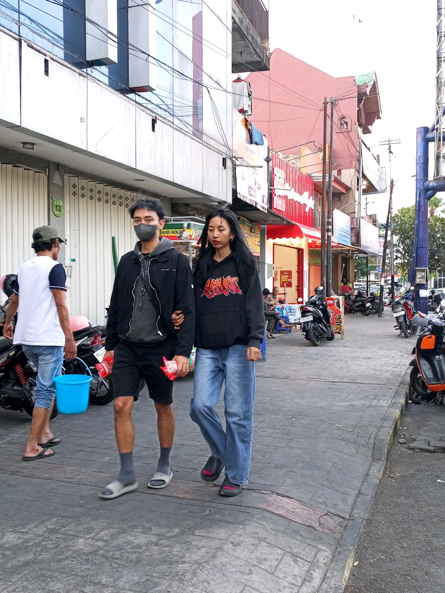 The image depicts a street scene in Batu, Indonesia. In the foreground, two young adults, a male and a female, are walking on a paved sidewalk. The male, wearing a black jacket, grey hoodie, black shorts, and grey socks with sandals, is walking on the left side of the female. He has a dark face mask covering his nose and mouth and is holding two red packets in his right hand. The female, wearing a black hoodie with a red graphic, blue jeans, and dark sandals, is walking on the right side of the male, holding his left arm. Behind them, on the left, another person wearing a white t-shirt with navy sleeves, blue jeans, and sandals is walking away from the camera, carrying a bright blue bucket. Several motorcycles are parked along the sidewalk and on the street. Buildings line the street, with some displaying commercial signage, including one with red corrugated metal siding. Overhead wires are visible, typical of urban infrastructure. The pavement is textured, and a slight incline is visible on the sidewalk. The lighting suggests it is daytime, possibly late afternoon, with soft shadows.