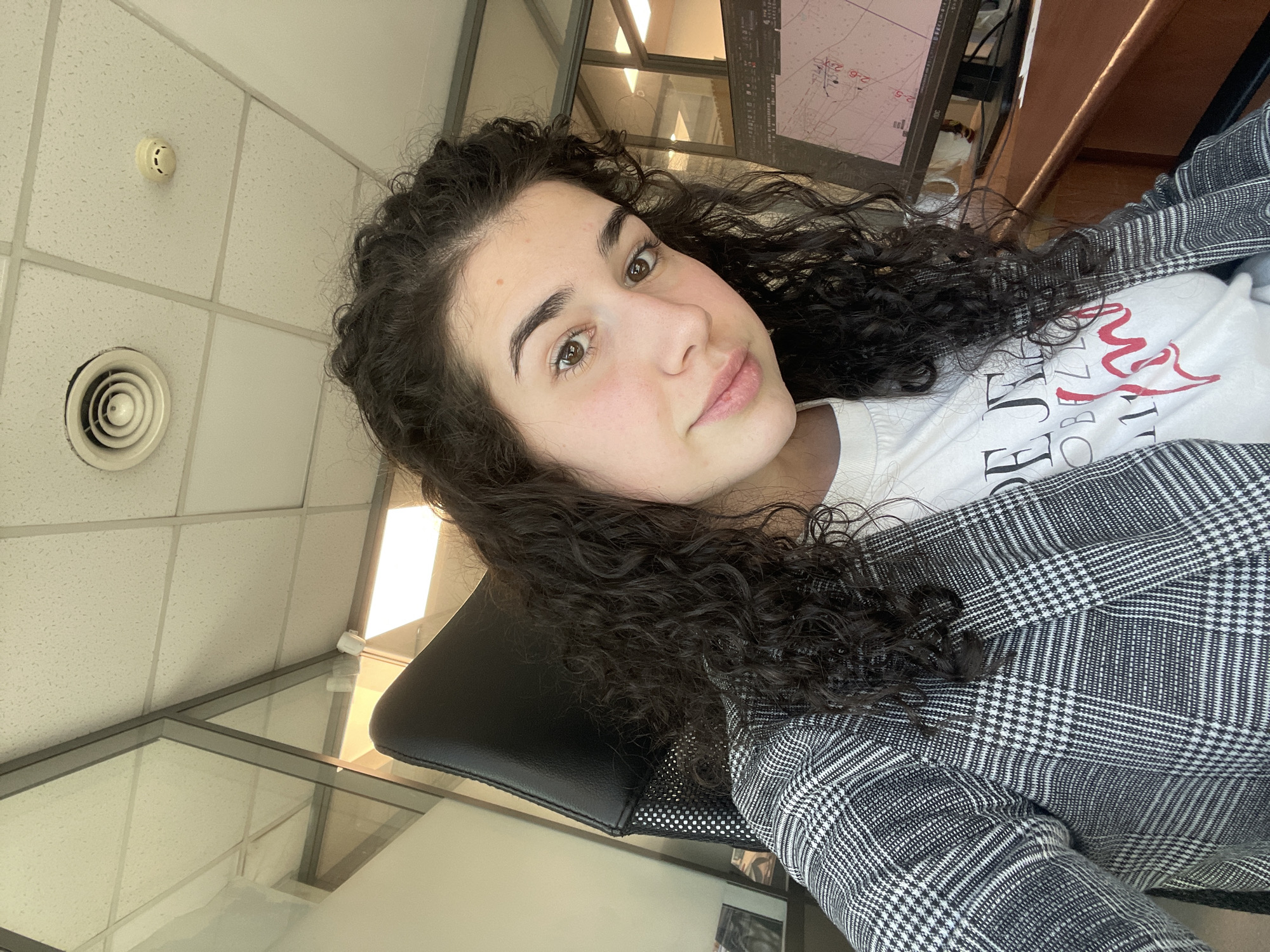 A woman with curly, dark hair is facing the camera with a slight smile. She has fair skin and wears a white T-shirt with red and black lettering under a gray and white plaid blazer. She sits in a black office chair. The background includes a standard office environment with ceiling tiles, an air vent, and a smoke detector. There is a glass partition and a desk with a computer monitor displaying a graphic interface. The exact location in Sofia, Bulgaria, cannot be determined from the available information.