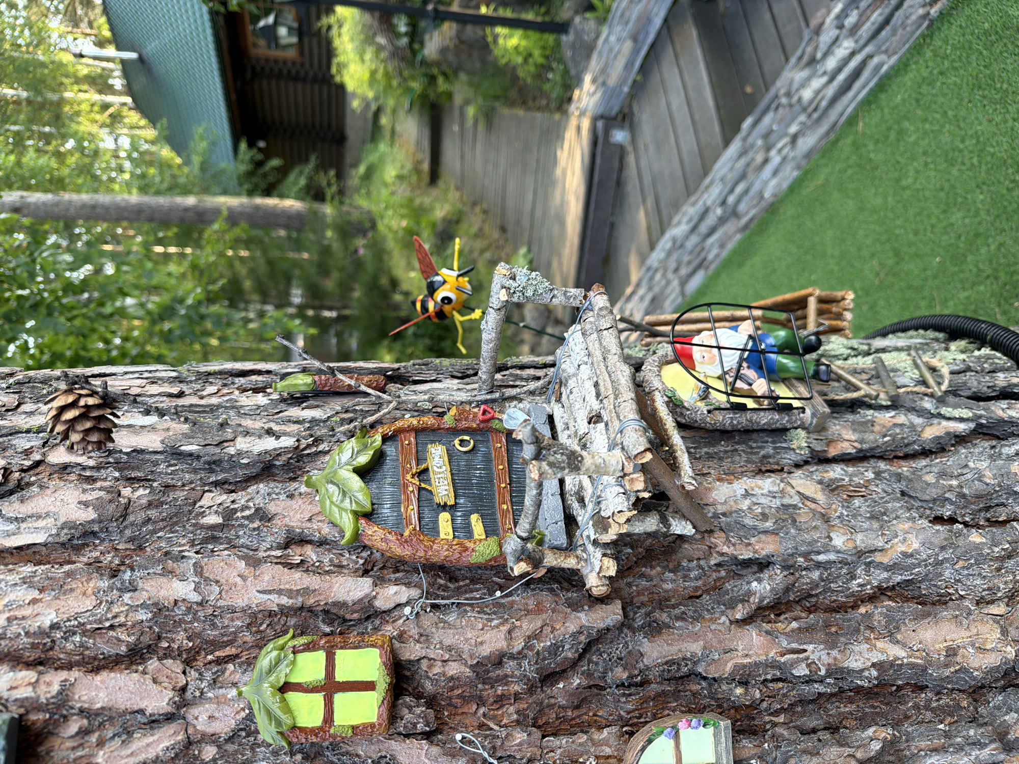This image depicts a detailed miniature fairy village attached to the trunk of a large tree. The village includes tiny houses, windows with green leaf-shaped frames, a "Welcome" sign, and a small wooden balcony. A gnome figure is visible inside a tiny enclosure, and a decorative bee figurine is positioned as if flying towards the village. The tree is set in a natural environment with a wooden pathway and a rustic log cabin visible in the background, suggesting a location in a rural or forested area. The presence of a green, artificial turf-like ground cover indicates a manicured garden setting. The overall scene is whimsical and intricately designed, creating a charming miniature world. The location in Sukhaya, Russia, implies a specific geographical context for this garden installation.