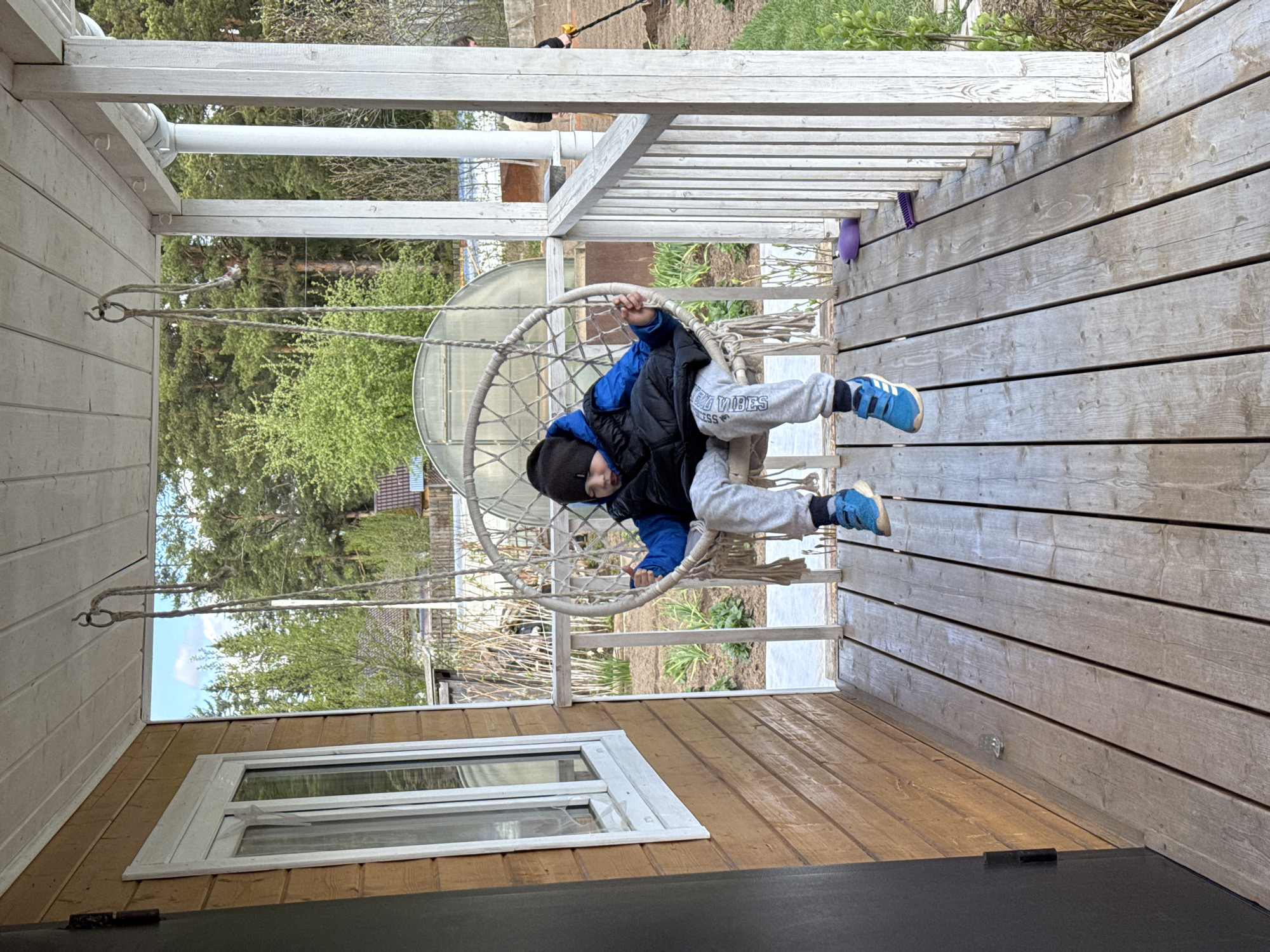 A child is sitting in a hanging chair on the porch of a wooden building. The child is wearing a black hat, a blue jacket, and gray sweatpants. The hanging chair is made of a white material and is suspended from the porch roof by ropes. The porch is made of light-colored wood and has a white railing. There is a window in the wall of the building to the left of the chair. Trees and a fence can be seen in the background. The image was taken in Chaikovsky, Russia.
