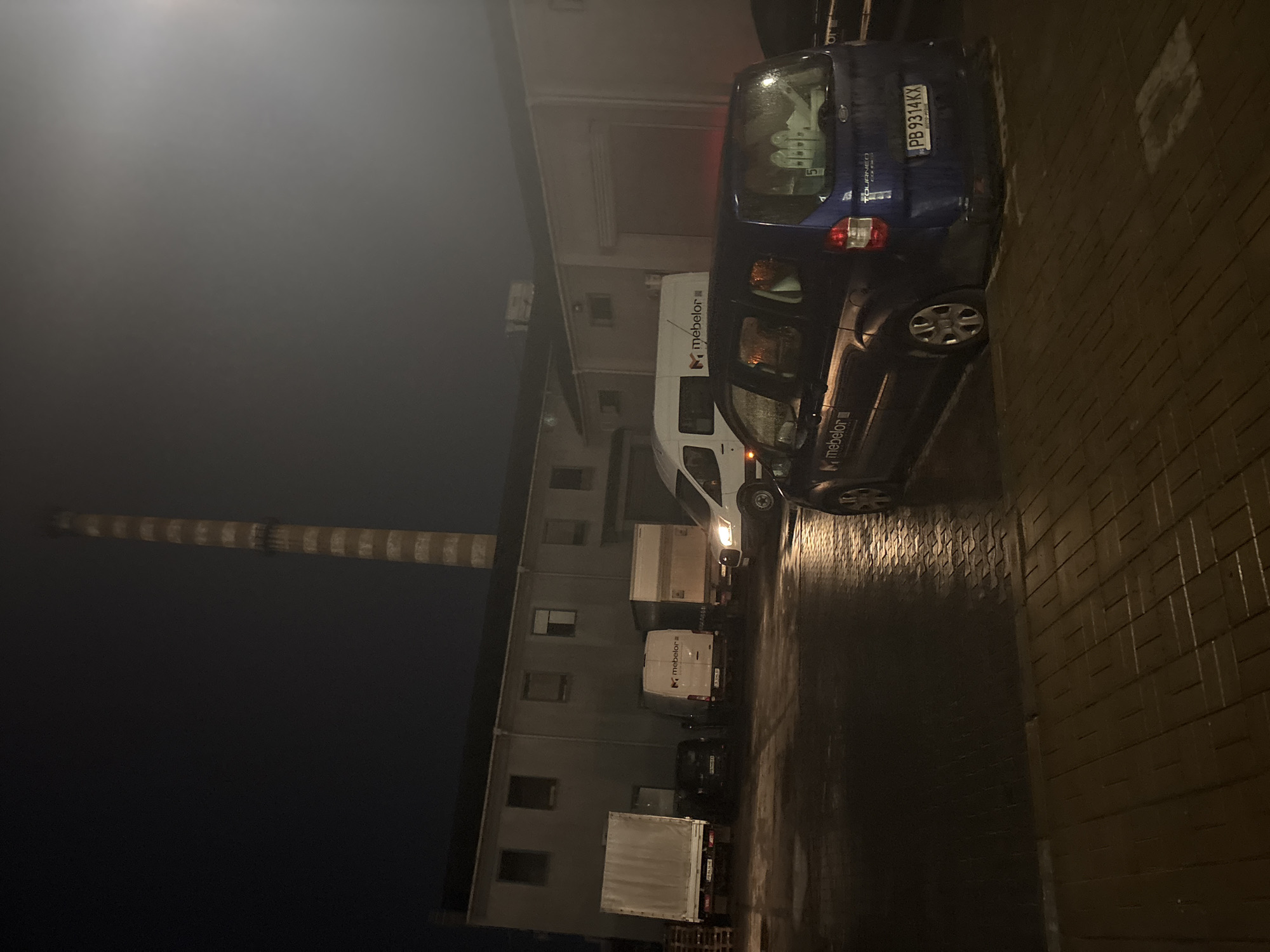 The image depicts a street scene in Plovdiv, Bulgaria, at night. The scene is dominated by vehicles parked along a building. A blue Ford Tourneo Connect with the license plate PB 9314 KX is parked closest to the camera. Several white vans with the "mebelor" logo are parked behind it. The building appears to be an industrial or commercial structure. A tall smokestack with alternating white and dark bands is visible in the background. The street is paved with dark bricks and wet, possibly from recent rain. The overall lighting is dim.