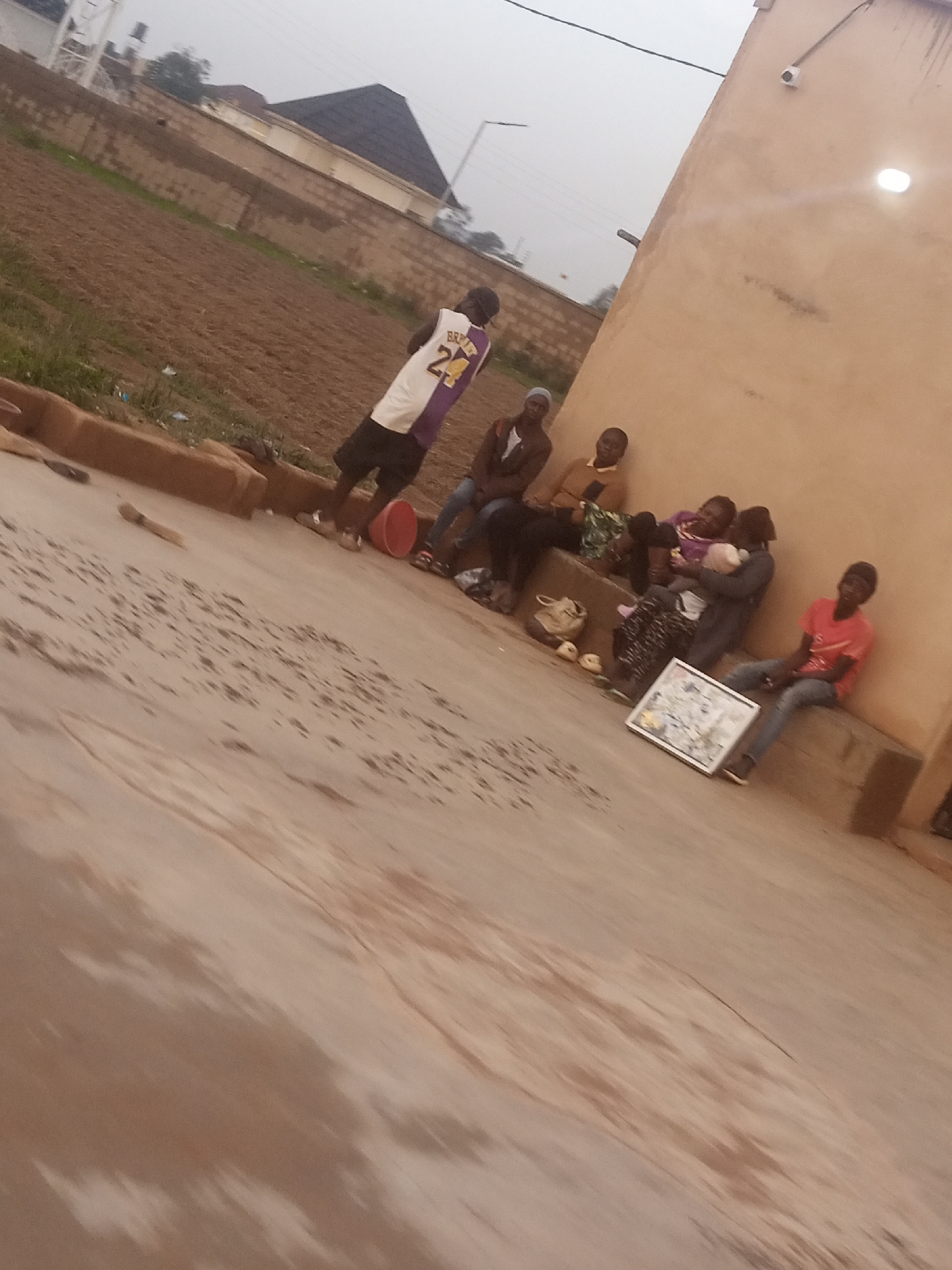 This media file shows a group of people gathered outdoors, possibly in Kwang, Nigeria. The scene appears to be a residential area with a building made of light-colored material, a dirt or field area, and a brick wall. The time of day seems to be overcast, possibly morning or late afternoon, due to the diffused light and muted colors.
The subjects include several individuals, some sitting and others standing. One person stands out wearing a purple and white basketball jersey with the number 24 on the back. Several people are seated on a raised concrete structure or bench in front of the building. Two individuals in the center appear to be holding and interacting with a baby. Another person is seated further to the right, wearing a red t-shirt. A framed picture or poster is propped up on the ground in front of the seated group. A red bucket and some bags are also visible on the ground.
The overall mood seems relaxed, with people gathered together, though it's difficult to discern specific emotions due to the image quality and distance. The setting suggests a casual outdoor gathering, perhaps a family or community get-together. The building has a security camera and a light fixture mounted on its exterior. The ground is a mix of paved and unpaved areas, with some visible puddles or damp patches. The distant background shows a dark-roofed structure and some vegetation.
