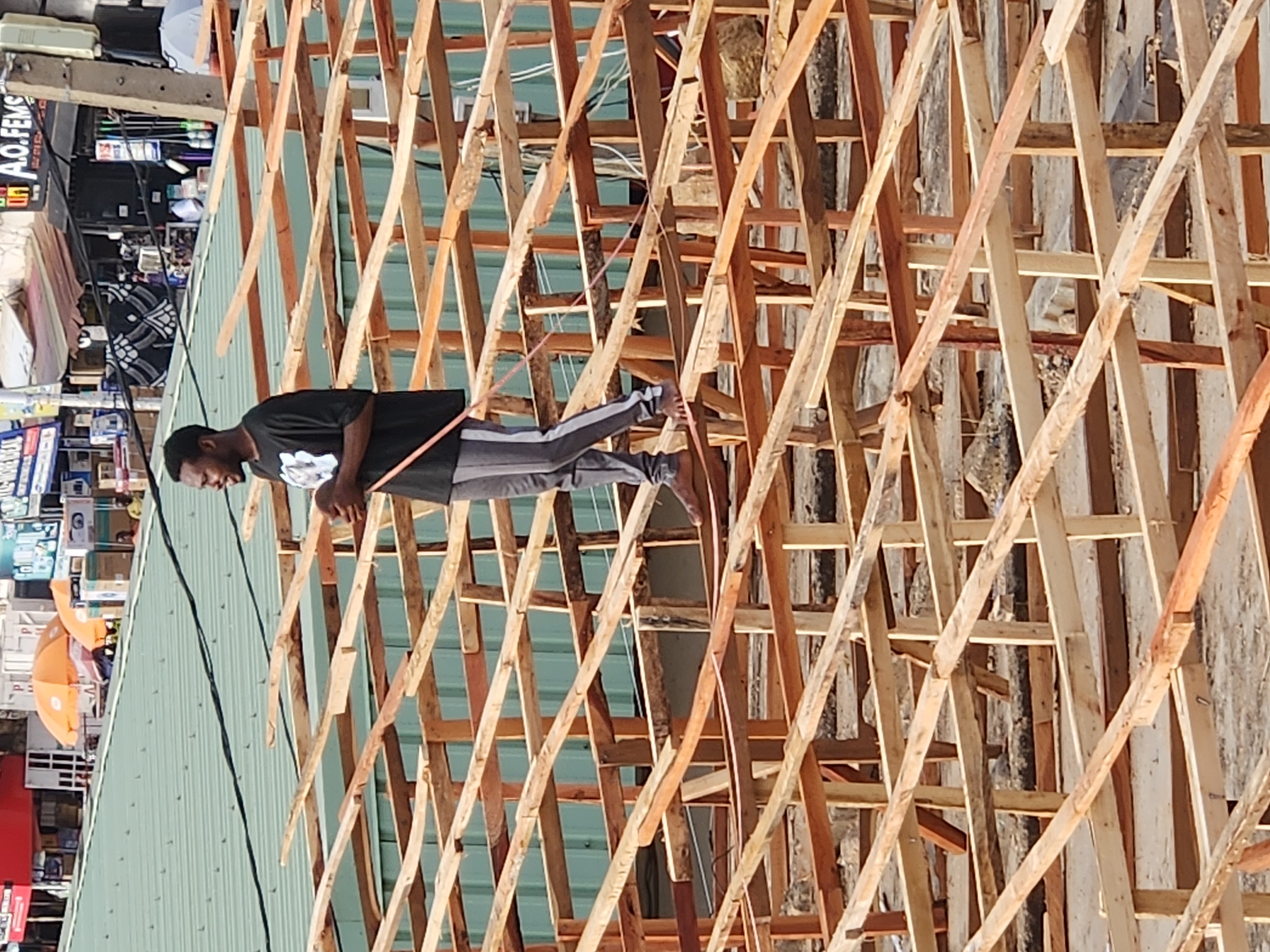A man stands on the wooden rafters of a roof under construction in Unknown, Unknown. He is wearing a dark t-shirt, grey striped sweatpants, and grey shoes. The roof is a latticework of light-colored wood. Below the roof, a green sheet metal roof is visible. Beyond that, storefronts and businesses line the street.