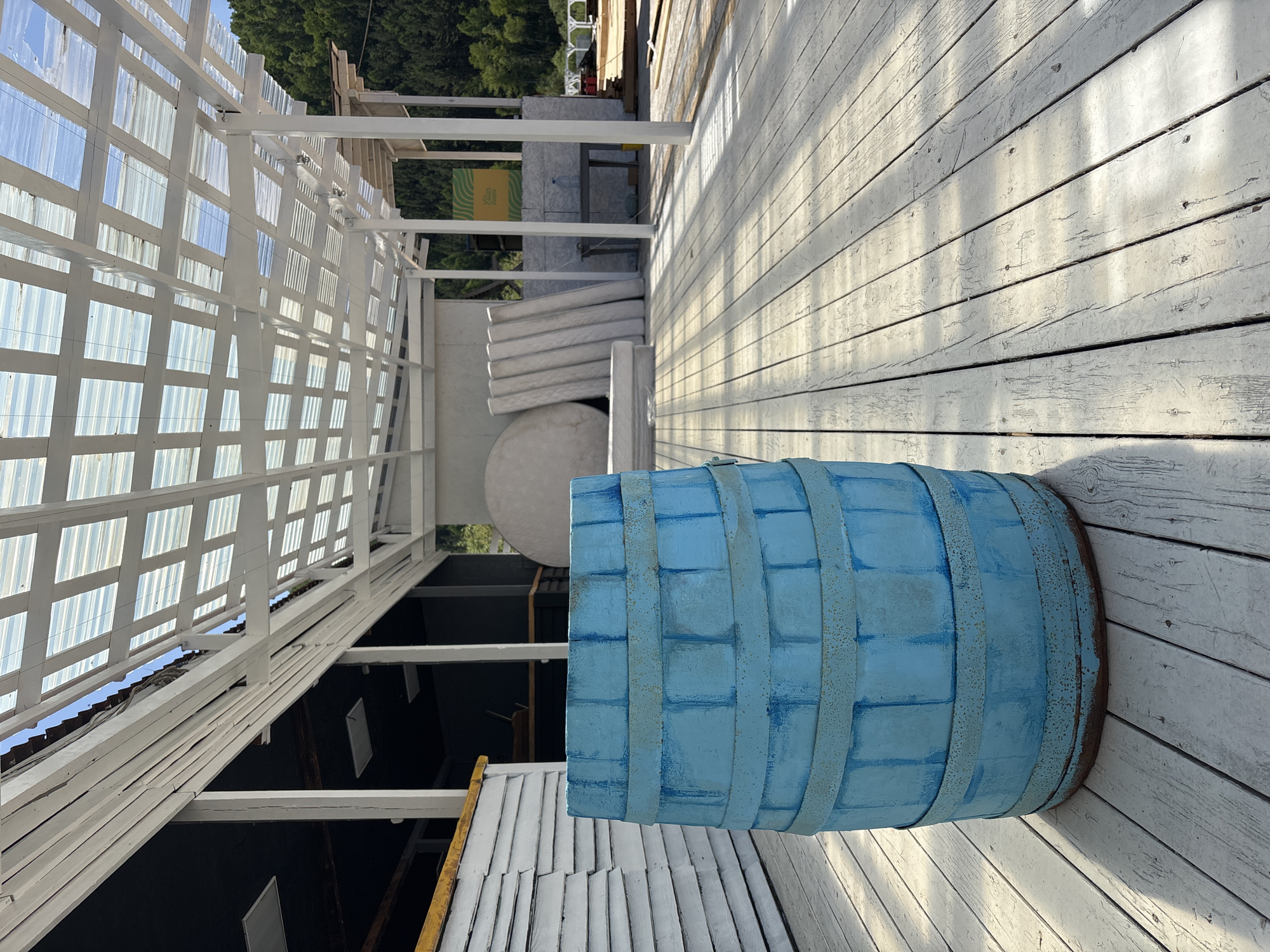 The image captures an outdoor leisure area, likely a terrace or deck, under bright daylight. In the immediate foreground, a large, light blue painted wooden barrel with darker blue accents and rusty metal bands rests on a white wooden plank deck. The deck planks are visibly aged with peeling white paint and some exposed wood grain, suggesting wear.
Overhead, a white wooden pergola structure with a translucent corrugated roof provides shade. Strong sunlight creates distinct shadows across the deck from the pergola's framework. In the mid-ground, further along the deck, several white, stacked cushions or mattresses are visible, along with what appears to be a large, light-colored spherical object. White support beams and posts frame the space.
Beyond the deck and structures, lush green trees are visible, indicating a natural, possibly forested, backdrop. The sky above is clear and blue. A yellow sign with green text and a wavy pattern is partially visible in the mid-ground; the text "VOLGA YOGA" and "ЙОГА НА ВЗЫМОСЛОВЕ" (Yoga on the hilltop/rise) can be discerned. The overall scene suggests a peaceful, outdoor recreational space, possibly part of a resort or wellness center in Дрокино, Russia. No people are present, and no specific activity is taking place, but the setting is prepared for relaxation or exercise.