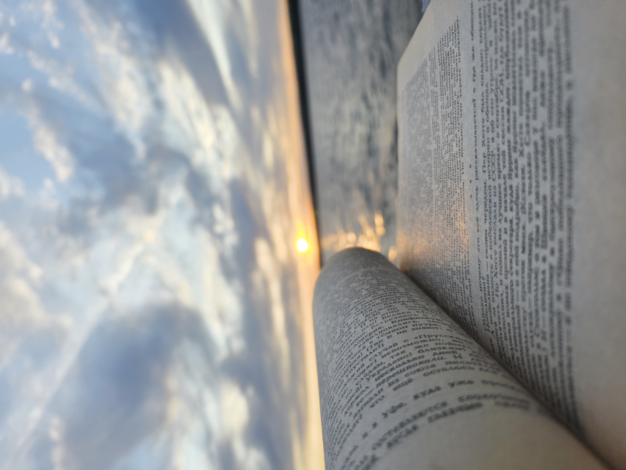 The image shows an open book with Russian text in the foreground. In the background, there's a blurred view of a sunset or sunrise over water, likely a river or a body of water, with clouds in the sky. The light from the sun is reflecting on the surface of the water. Given the presence of Russian text, the image might be taken in Russia.
The information is not specific enough to confirm that it was taken in Chaikovsky, Russia.