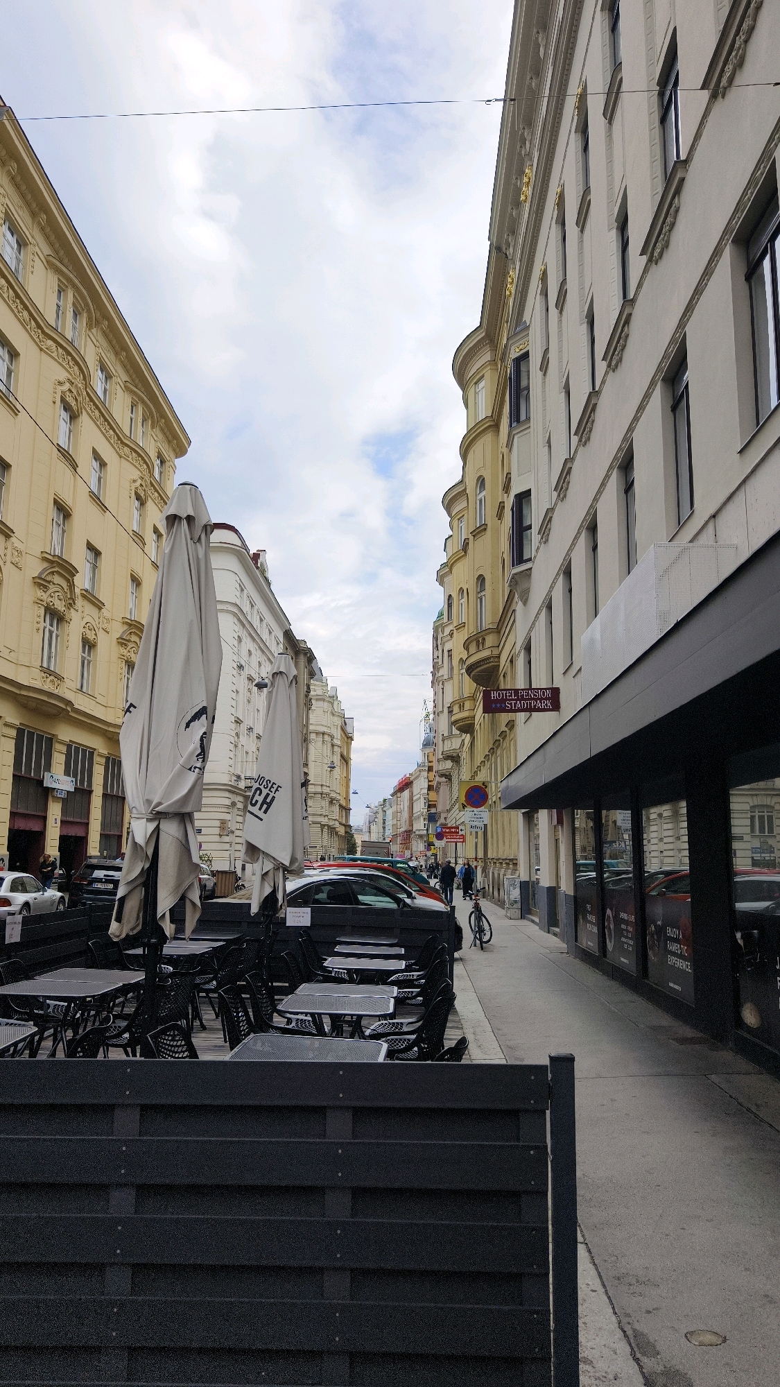 The image shows a street scene in Vienna, Austria. On the left side of the street, there are several buildings with yellow facades and ornate architectural details. In front of these buildings, there is an outdoor seating area with black tables and chairs, partially covered by large, closed white umbrellas. Cars are parked along the curb.
On the right side of the street, there is a building with a white facade and dark windows. A sign on this building reads "HOTEL PENSION STADTPARK." The sidewalk in front of this building is lined with more cars and some pedestrians. A bicycle is parked on the sidewalk. The sky is cloudy.
