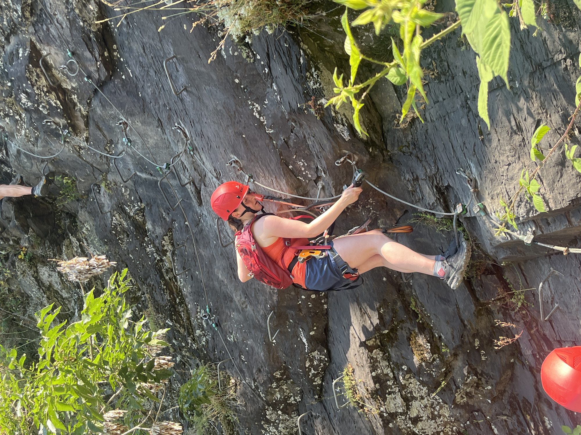 A person wearing a red helmet, orange tank top, and blue shorts is rock climbing on a steep, rocky cliff face. They are secured to a safety cable with carabiners and appear to be in the middle of traversing the route. Green foliage grows from the cliff and a red climbing helmet is visible in the lower right corner of the image. The sun is shining, creating shadows on the rock, suggesting it is daytime. The overall scene depicts an outdoor adventure activity.