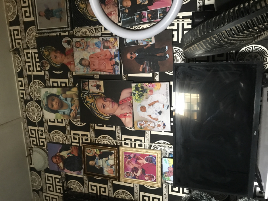 The image displays an interior wall within a residence in Oba-Ile, Nigeria, densely covered with various framed and unframed photographic prints and canvases. The wall is adorned with black and white wallpaper featuring a repeating geometric pattern composed of interlocking lines and circular medallions.
Numerous portraits of individuals are mounted on the wall. These include multiple adult females, often depicted wearing traditional Nigerian headgear (gele) and beaded necklaces, some of these prints incorporating smaller inset photographs of children or alternative poses. Portraits of adult males and numerous children and babies are also visible. Specific examples include a baby in a white outfit with "Abdul Hafeez @ 3months" inscribed below, a child in traditional attire, and a child wearing a suit and sunglasses. Several photographs are enclosed in ornate gold-colored frames, while others are canvas prints. A light-colored, semi-circular decorative object is affixed to the upper left portion of the wall, and a horizontal wire with small clips is suspended across the upper wall section.
Positioned beneath the wall display is a black LG flat-screen television, whose screen exhibits smudges and reflects an overhead light source or window. To the right of the television, the gridded, dusty back of a black electric fan is partially visible, angled upwards. A green, tinsel-like decorative element is partially observable to the left of the television.