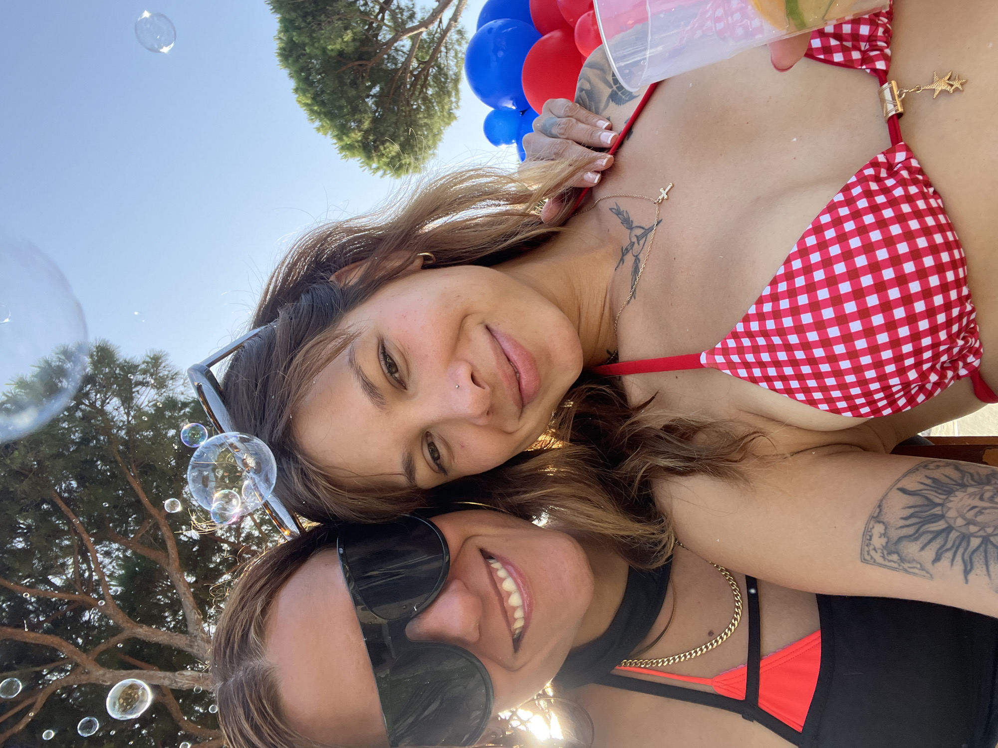 In the city of Sant Vicenç De Montalt, Spain, two women are taking a selfie outdoors. The woman on the left wears large black sunglasses, a black top with orange trim, and a gold chain. She is smiling. The woman on the right, also smiling, wears a red and white gingham bikini top and a gold cross necklace. She has tattoos on her arm. In the background, there are trees and a clear blue sky. Several bubbles are visible in the air, and some large blue and red balloons are clustered behind the woman on the right. One woman is holding a clear plastic cup with a drink. The overall scene suggests a casual, festive outdoor gathering.