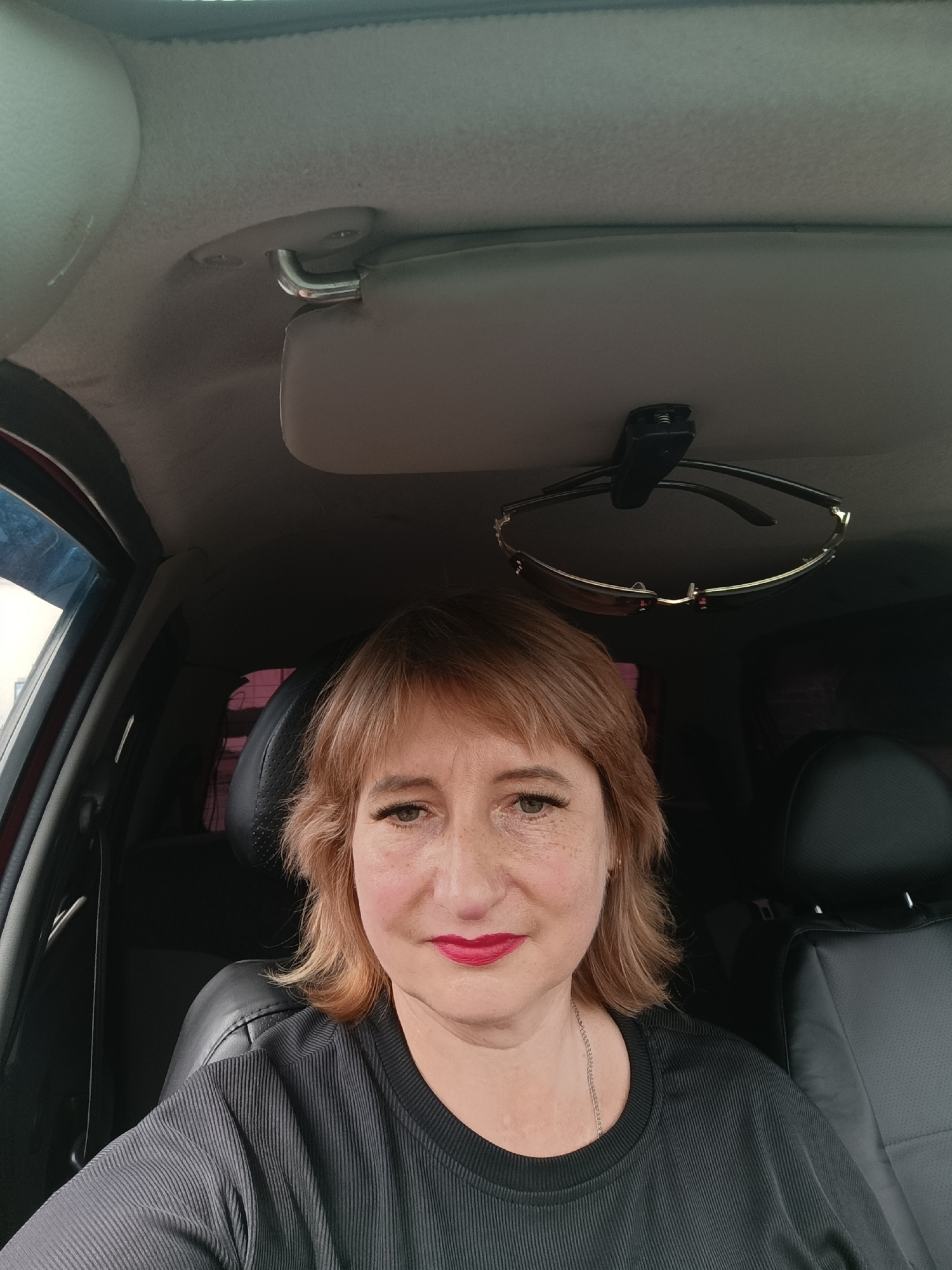 A medium shot captures a middle-aged Caucasian woman with auburn hair and light skin, wearing bright red lipstick and a black, ribbed shirt, seated in the driver's seat of a car. She has freckles scattered across her nose and cheeks and wears eyeliner and mascara. Her left eye is open and looking at the camera, while her right eye is partially visible. A pair of sunglasses hangs from a clip on the car's ceiling, just above her head. The interior of the car is visible, with black leather seats and the car's headliner. The lighting suggests it is daytime, with natural light illuminating the scene from the front and left. The image appears to have been taken inside a vehicle.